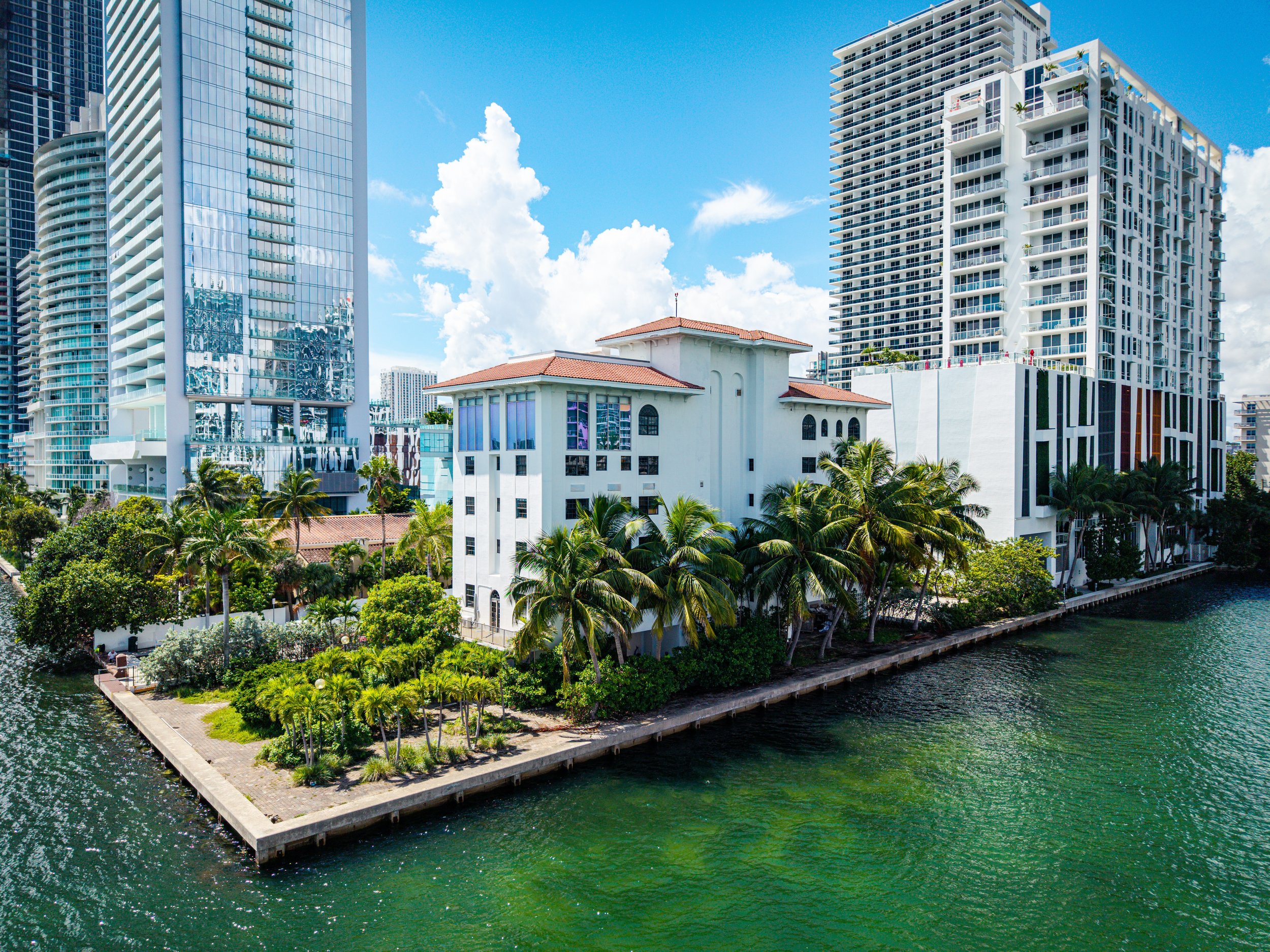 A cityscape with tall modern buildings alongside a waterway, with palm trees and a small pier or walkway in the foreground. The sky is blue with white clouds.