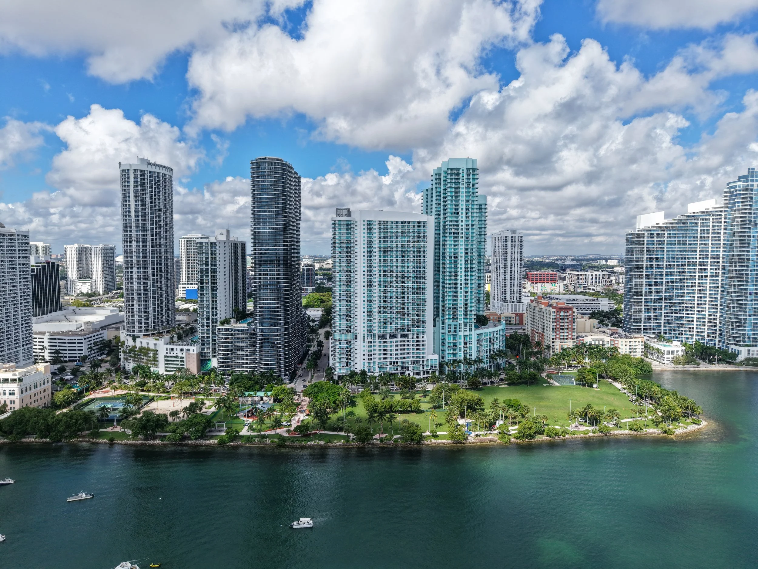 A city skyline with tall modern high-rise buildings next to water and a park with green trees and grass, under a partly cloudy sky.