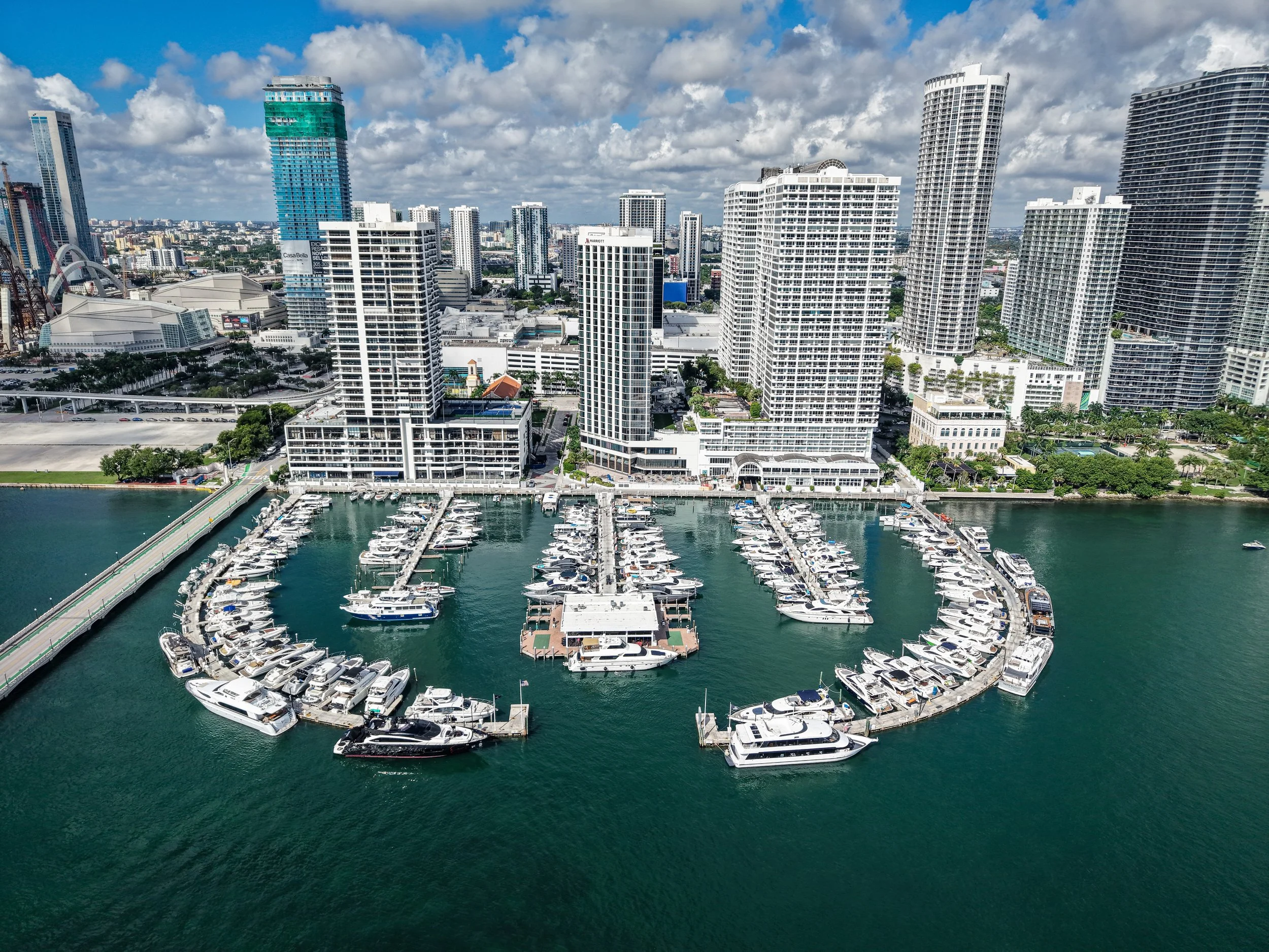 Aerial view of Miami skyline with numerous high-rise buildings and luxury yachts docked at a marina along the waterfront.