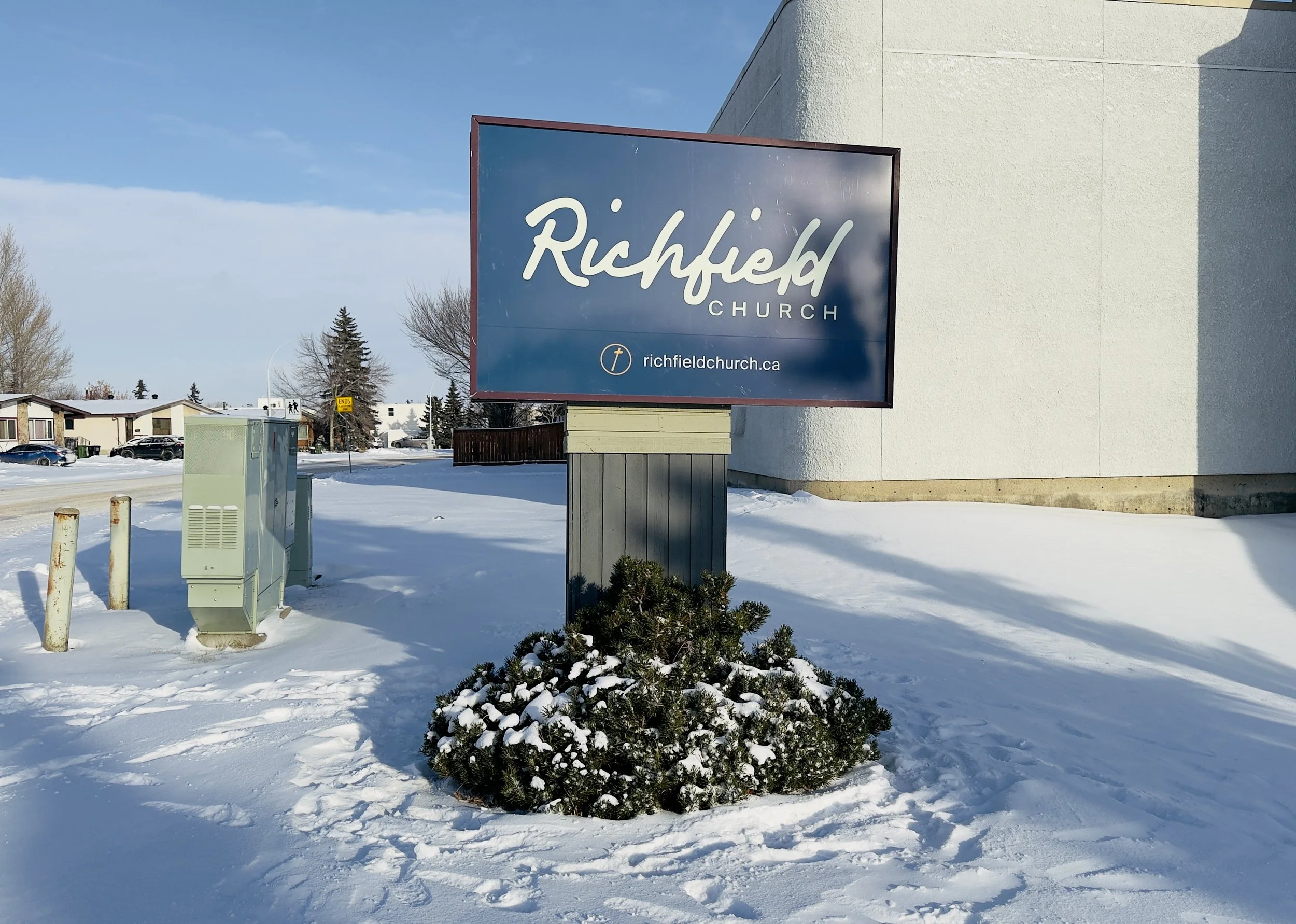 Church sign for Richfield Church with website URL richfieldchurch.ca, in a snowy outdoor setting with a small bush, utility boxes, and nearby residential buildings.