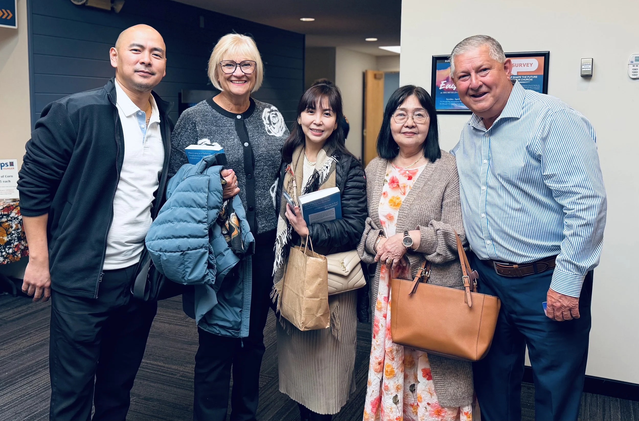 Group of five people smiling and standing together indoors, holding books and bags.