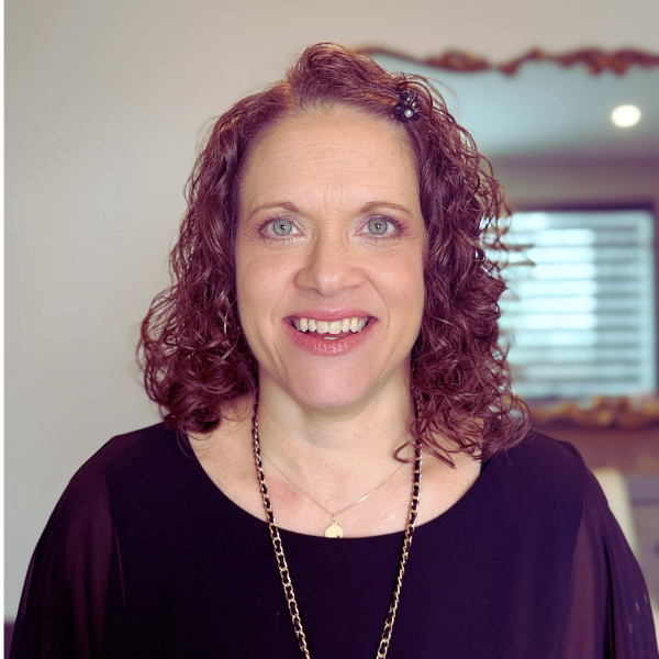 A woman with curly reddish-brown hair and blue eyes smiling at the camera, wearing a black top and a gold necklace, in a room with a mirror and window in the background.