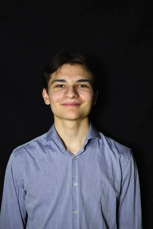 Ben smiling, wearing a light purple button-up shirt, standing against a black background.