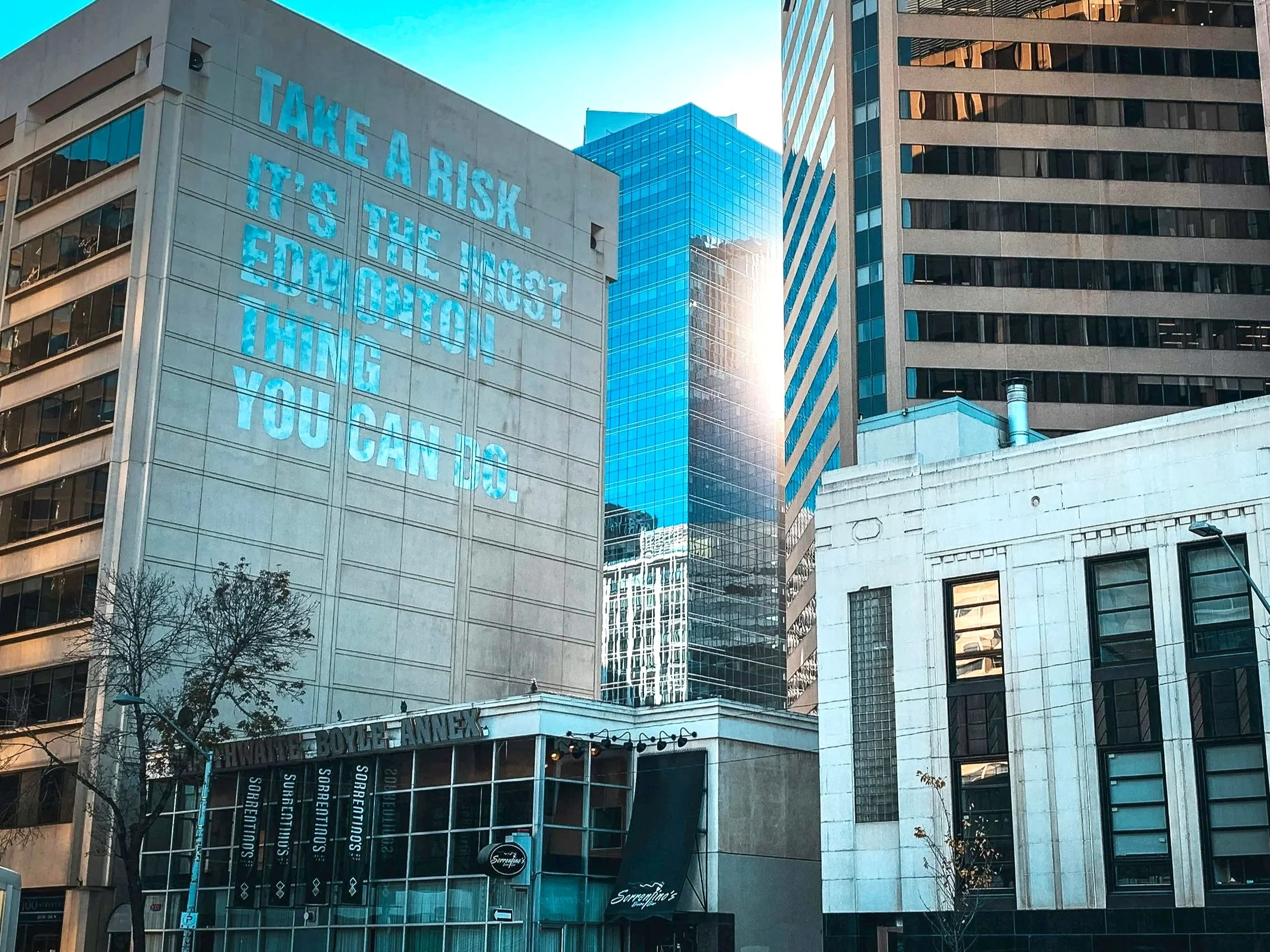 Multiple tall buildings in a city with a blue sky. One building has a large digital sign displaying a motivational quote about taking risks and believing in oneself.