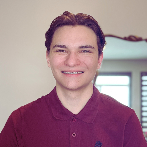 Smiling young man with short brown hair wearing a maroon polo shirt, standing indoors near a window.