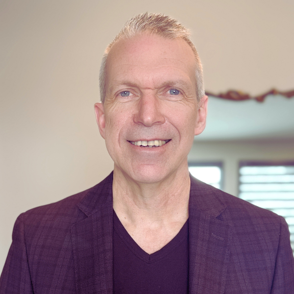 A middle-aged man with short, light-colored hair is smiling, wearing a dark blazer over a black shirt, standing indoors with windows in the background.