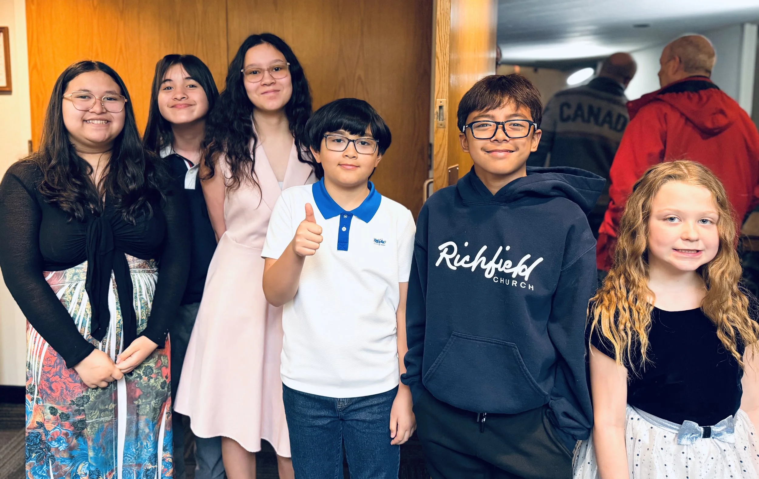 A group of six children standing together and smiling for the photo. One boy is giving a thumbs-up. The children are dressed casually, and some are wearing glasses. Two adults are visible in the background, talking to each other.