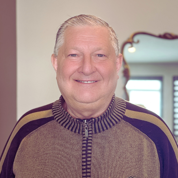 A smiling middle-aged man with short gray hair standing indoors with a mirror and window in the background.