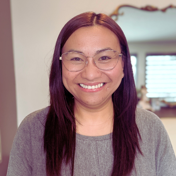 A woman with long, straight purple hair wearing glasses and a gray top, smiling indoors.