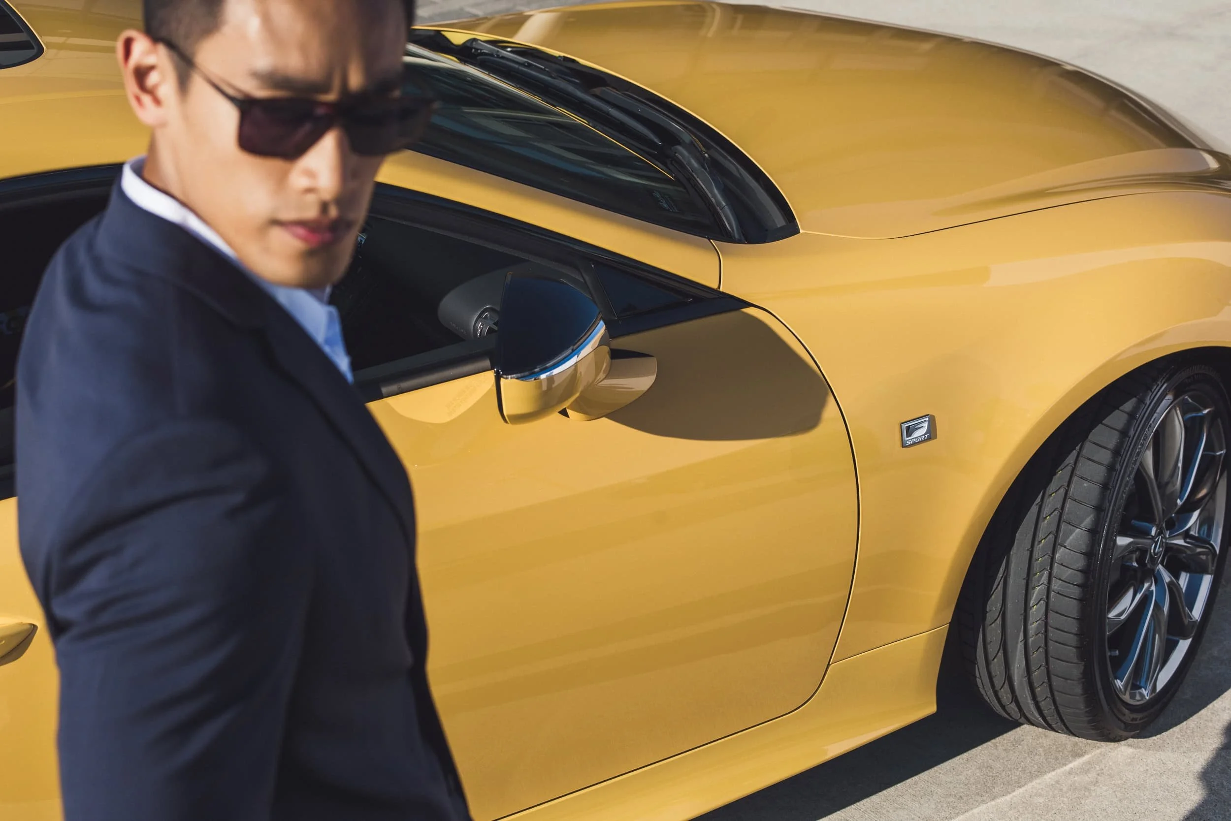 A man in dark sunglasses and a dark jacket standing in front of a yellow sports car with black wheels, side mirror, and a general sleek design.
