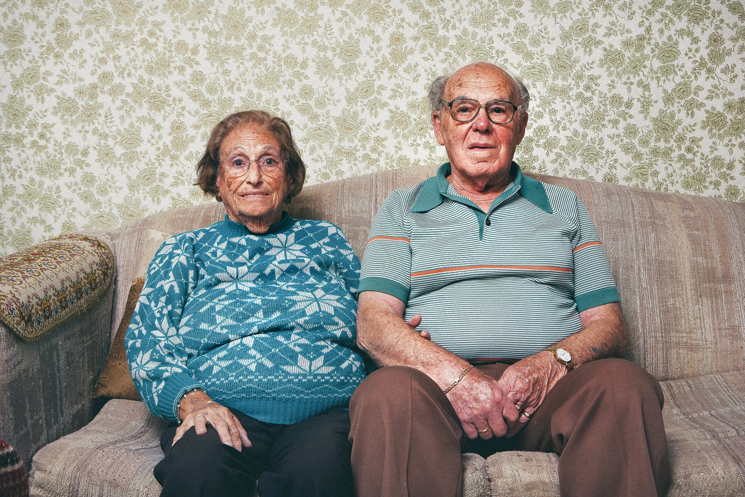 An elderly couple sitting on a old patterned sofa in a room with floral wallpaper. The woman is wearing a blue sweater with white snowflakes and the man is wearing a striped polo shirt. Both are wearing glasses.