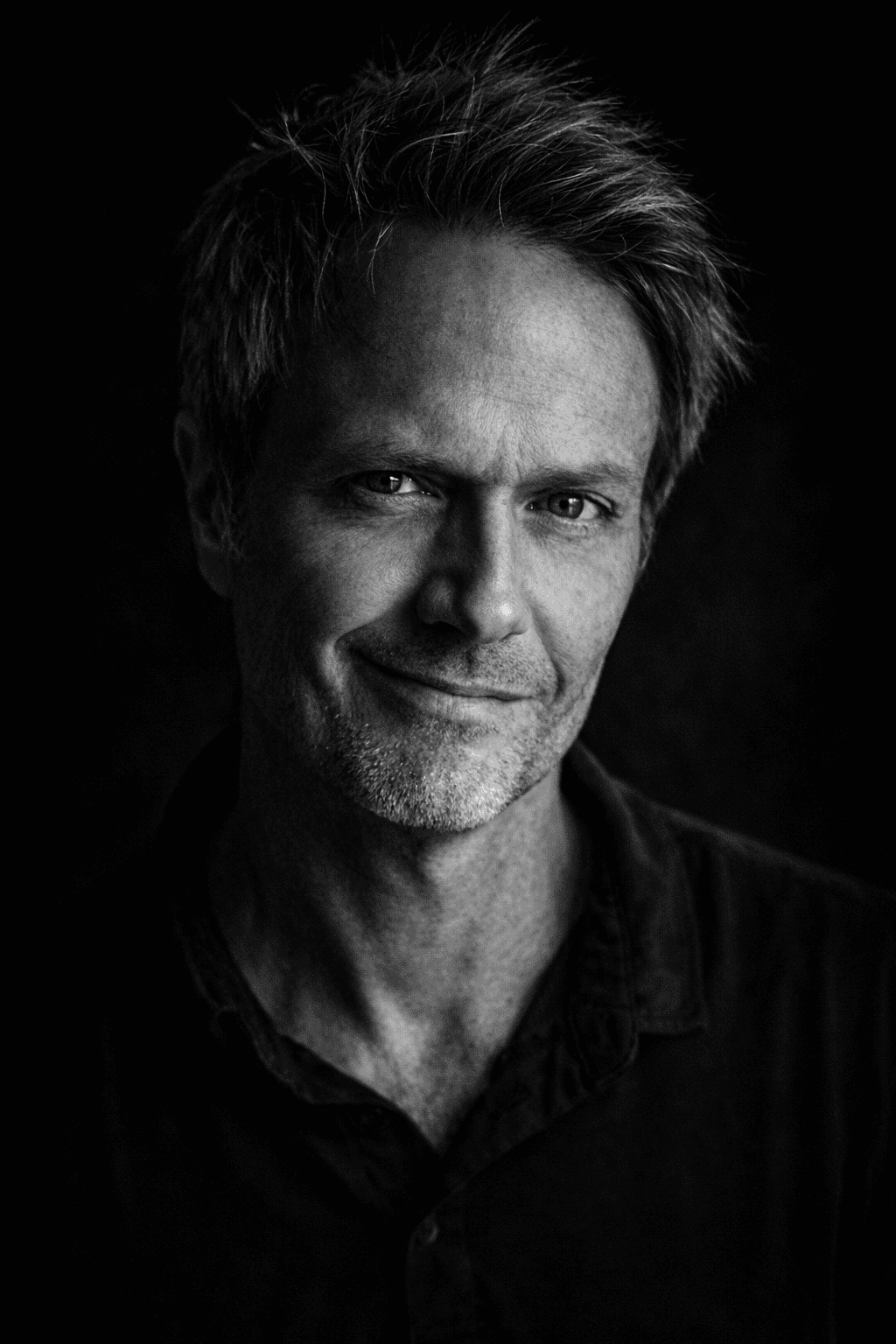 Black and white portrait of a middle-aged man with short, tousled hair, smiling slightly, wearing a dark shirt, dark background.