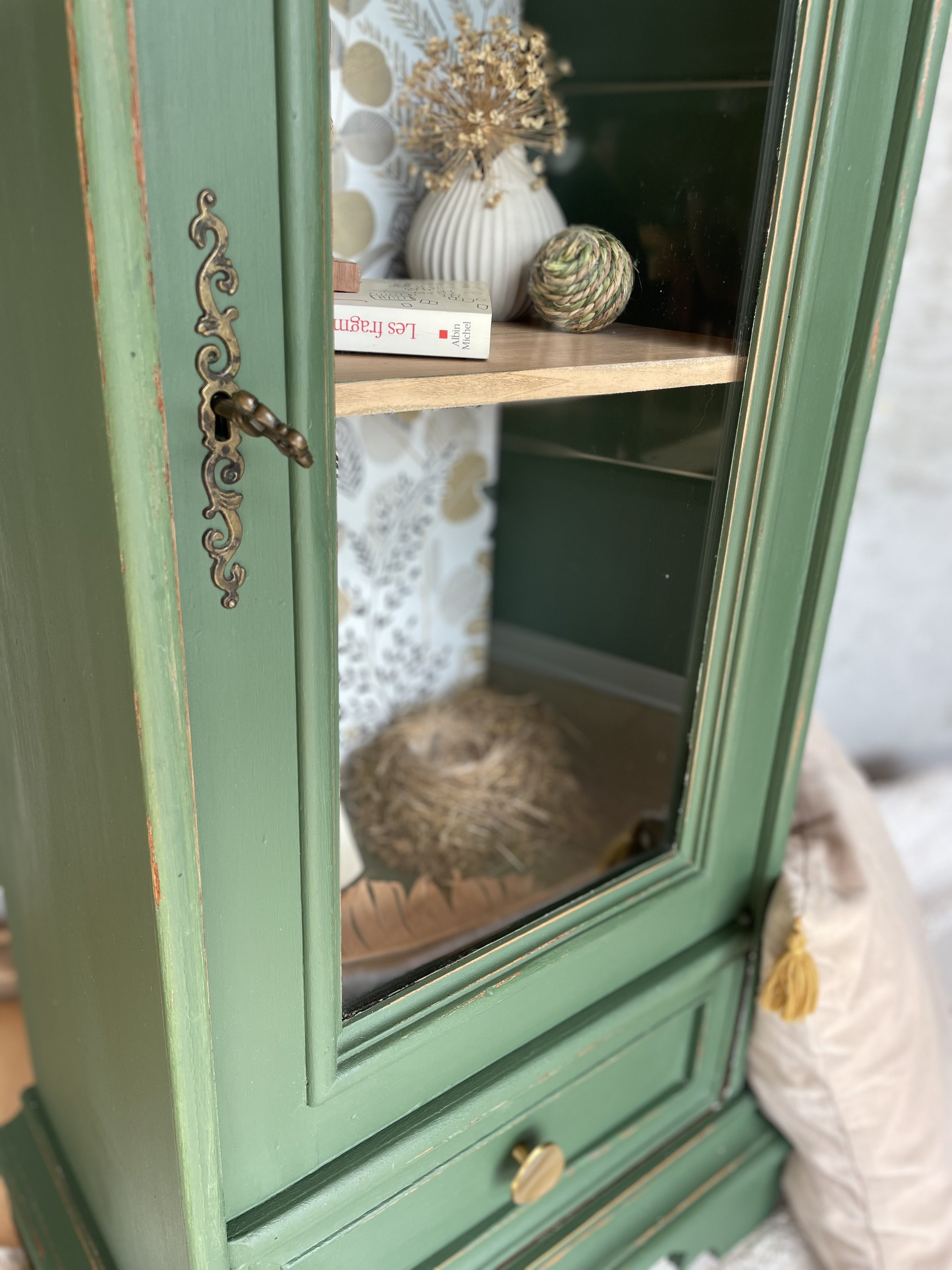 Une armoire en bois peinte en vert avec un verre en façade, contenant des décorations comme un vase blanc avec des branches séchées, une boule en rotin, et un livre. La pièce en arrière-plan montre un mur avec un motif floral.