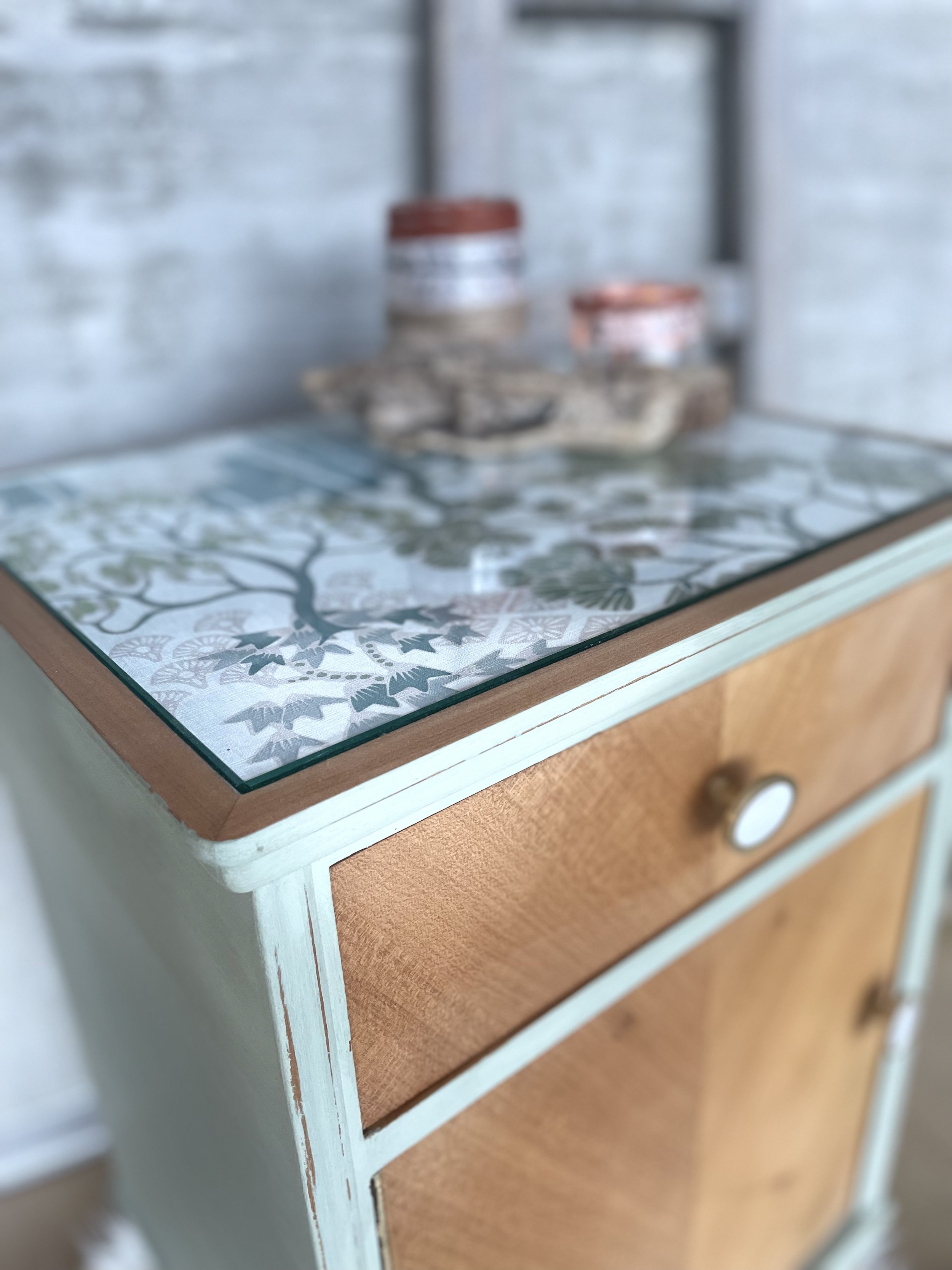 Une petite commode en bois avec un dessus en verre décoratif. En arrière-plan, des bocaux en conserve ou en épices sont visibles.