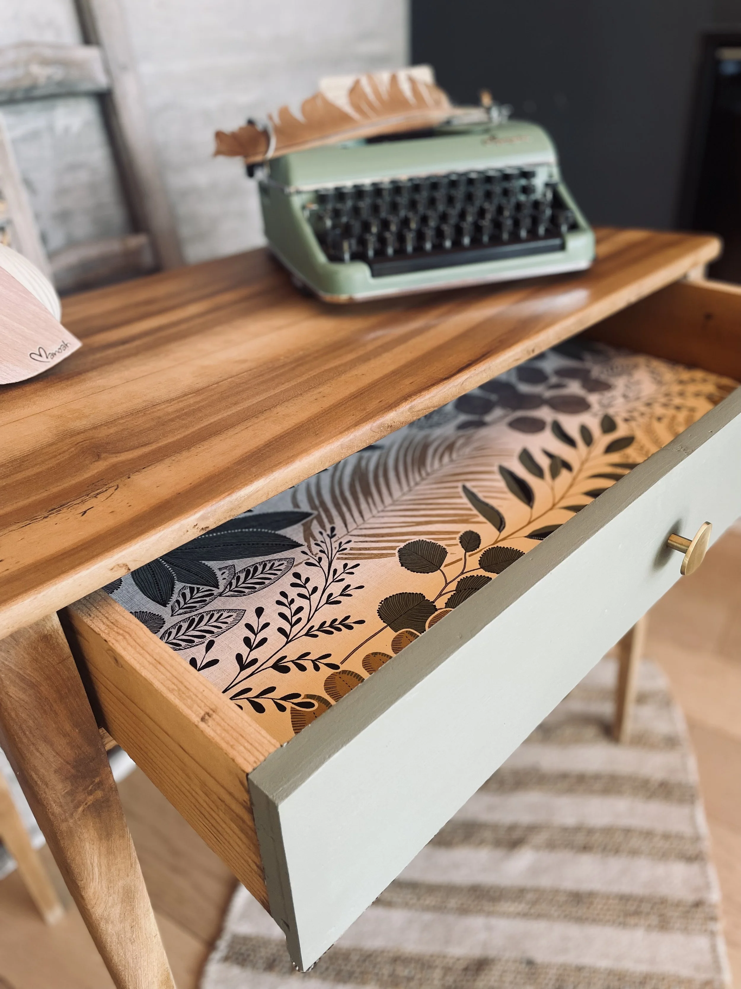 Un bureau en bois avec une vieille machine à écrire verte posée dessus, et une tiroire partiellement ouverte avec un intérieur décoré de motifs floraux en noir et beige.