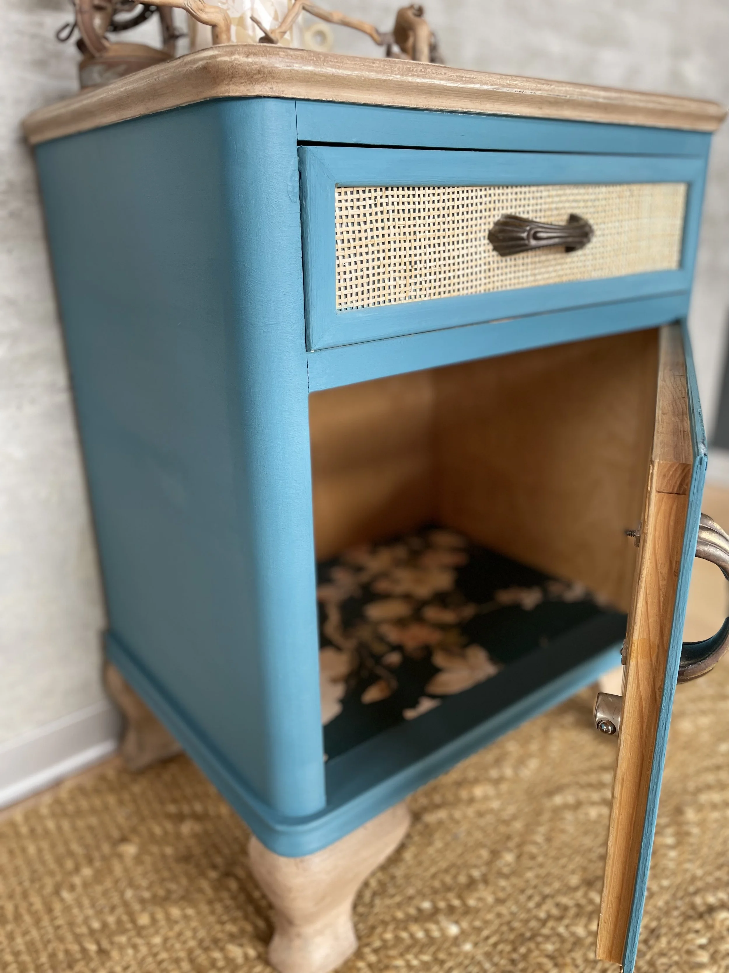 Une petite armoire bleue avec un tiroir à motif en osier et un tiroir en bois massif, avec une porte ouverte révélant l'intérieur en bois et un fond décoré de feuilles. La pièce est posée sur un tapis en jute.