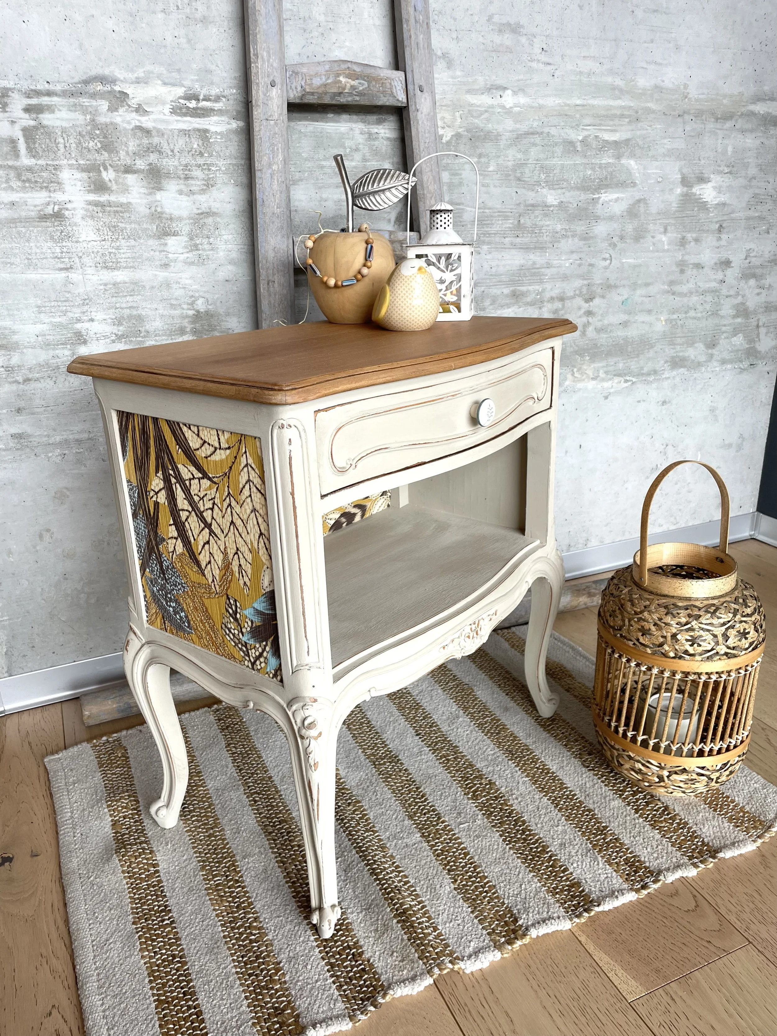 Une console blanche avec une surface en bois clair décorée d'objets comme un pot en forme de citrouille, une petite figurine en forme de chouette, une lanterne blanche et un collier de perles. La console repose sur un tapis rayé beige et blanc, à côté d'un panier en rotin avec un motif crocheté.