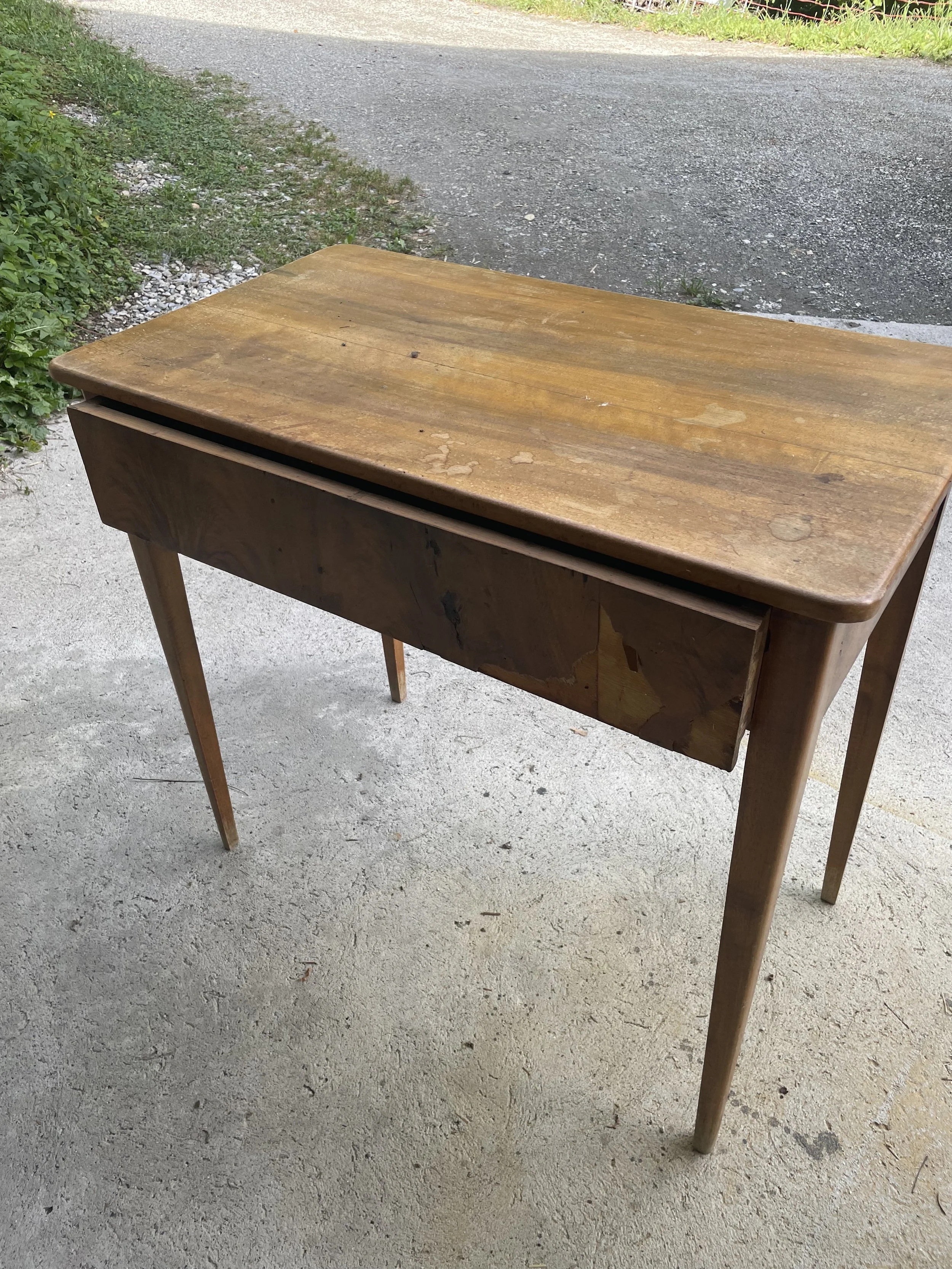 Table en bois usée et abîmée située à l'extérieur sur un sol en béton, avec de la végétation en arrière-plan.