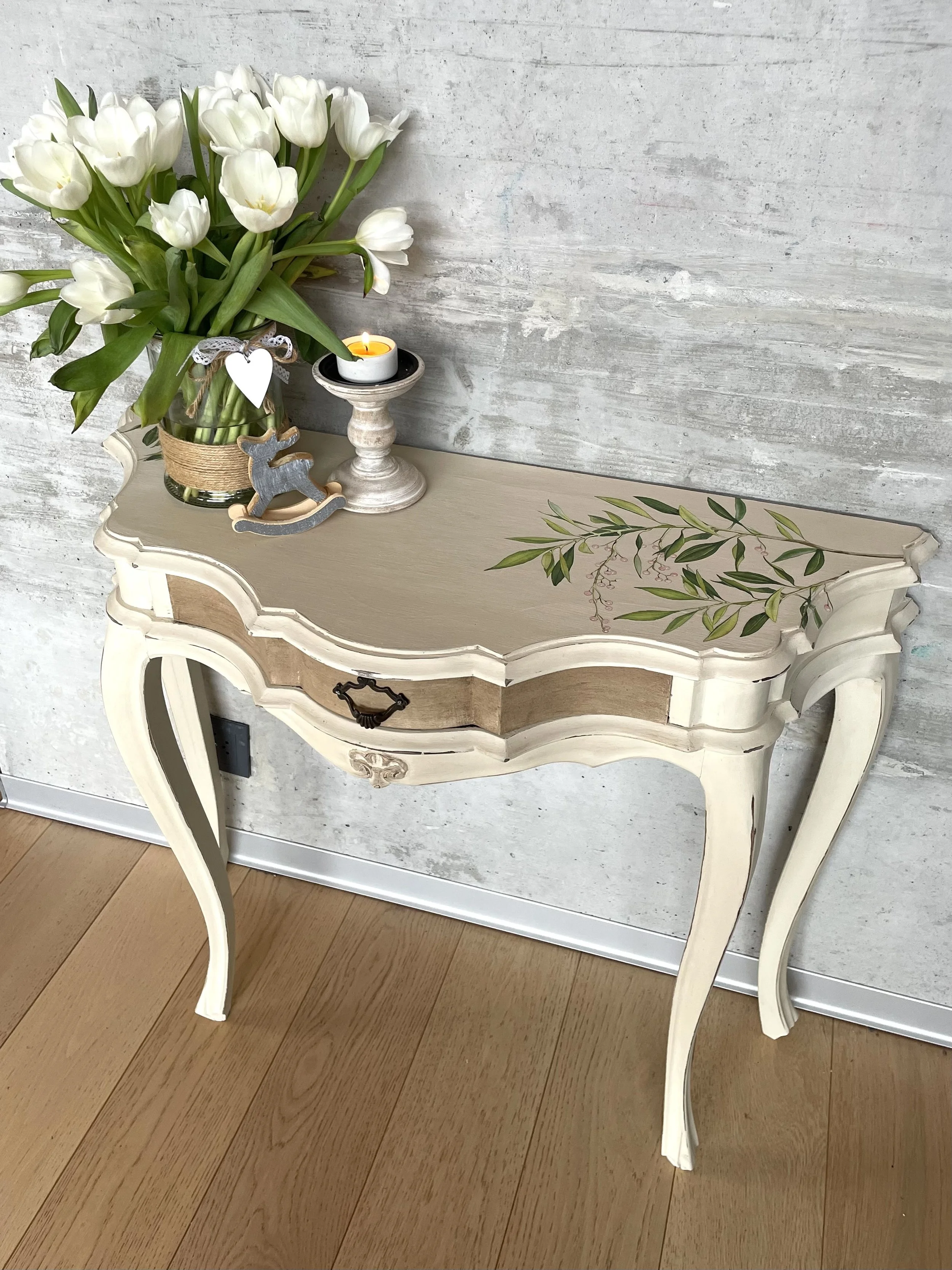 Une petite table ancienne de style shabby chic avec un plateau beige clair, une poignée en métal noir, et des pieds courbés, décorée d'un bouquet de tulipes blanches dans un vase, d'une bougie allumée dans un support en bois, et d'un petit rocking horse en bois gris.