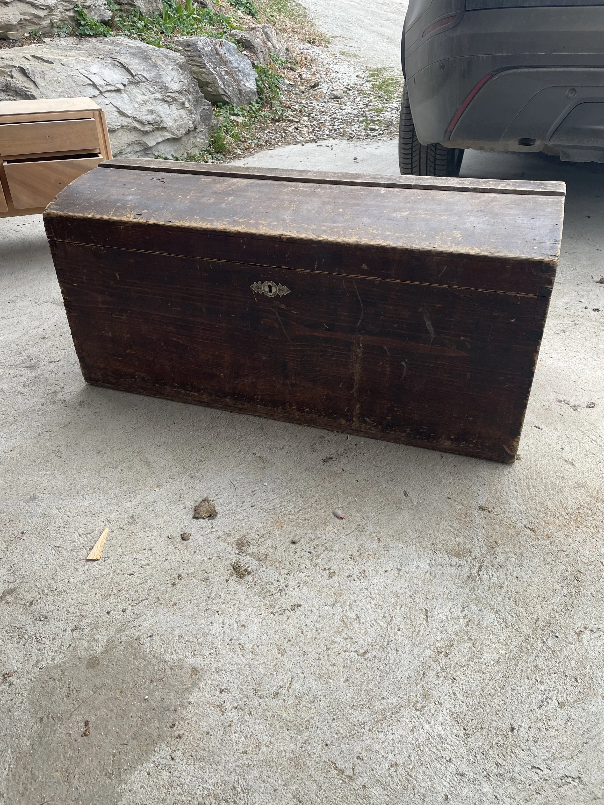Un coffre en bois ancien, sombre, placé sur un sol en béton, avec une voiture en arrière-plan et des rochers et des plantes à proximité.
