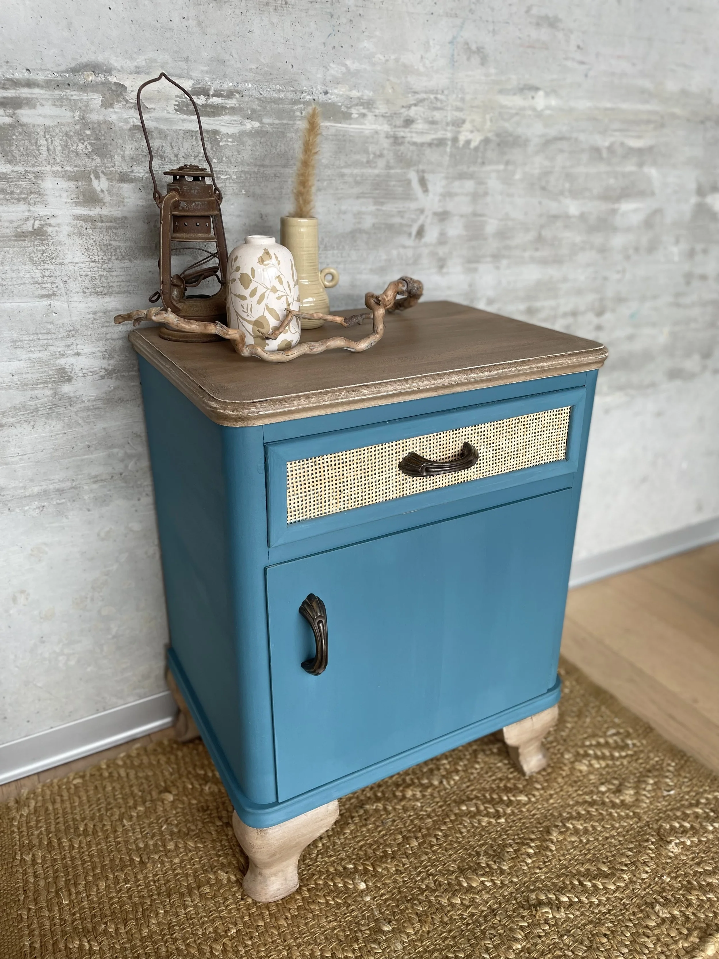Une petite armoire bleue avec un dessus en bois clair, décorée avec des objets en terre cuite, une lanterne ancienne, un vase, et une branche décorative, devant un mur en béton.