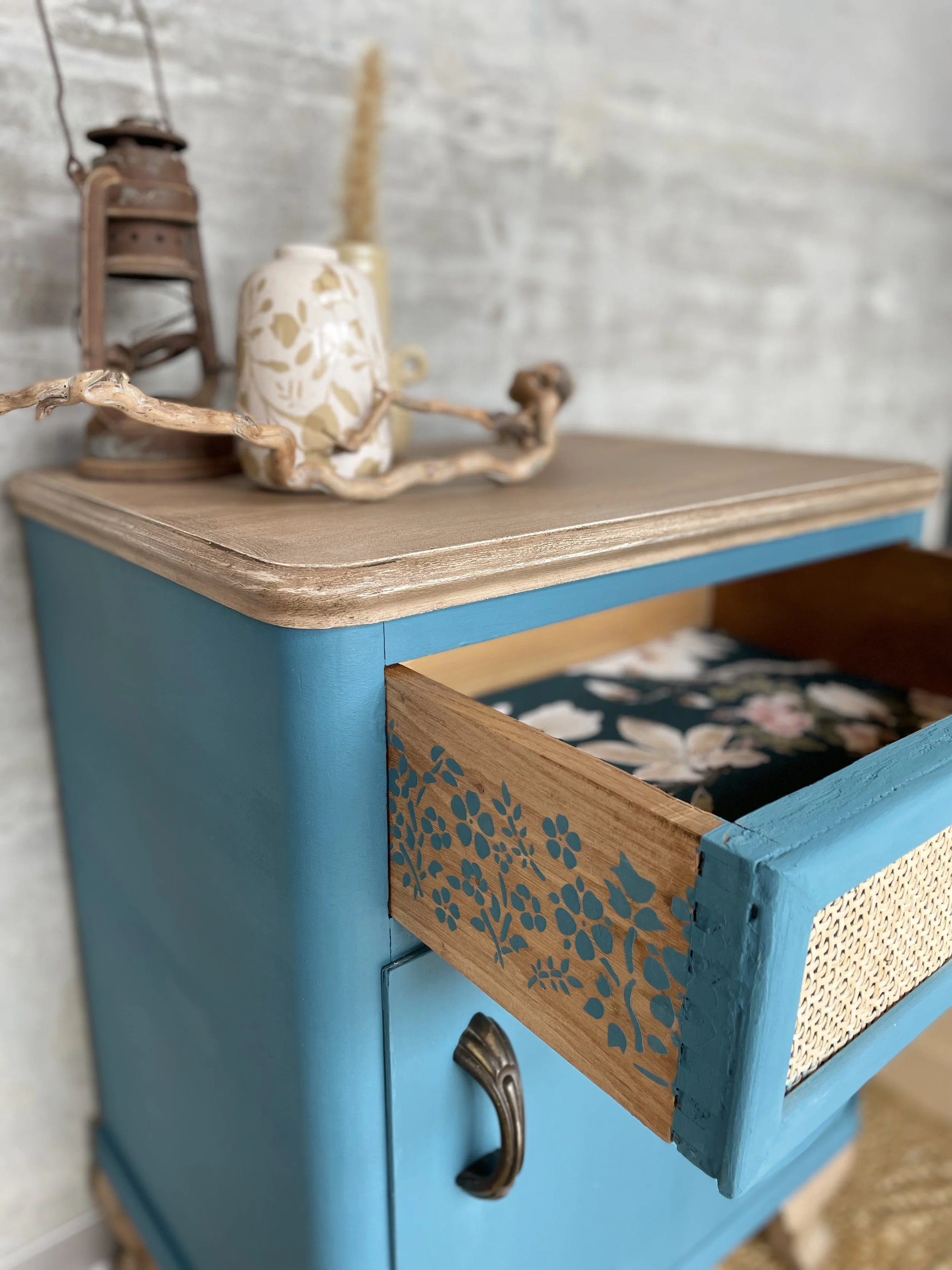 Un meuble turquoise avec une porte en bois peint avec un motif floral bleu, une poignée noire, et un tiroir partiellement ouvert révélant un intérieur tapissé de tissu floral. Sur le dessus en bois clair, il y a une vieille lanterne en métal rouillé, une céramique blanche avec des motifs dorés, un morceau de bois torsadé et un fond gris clair
