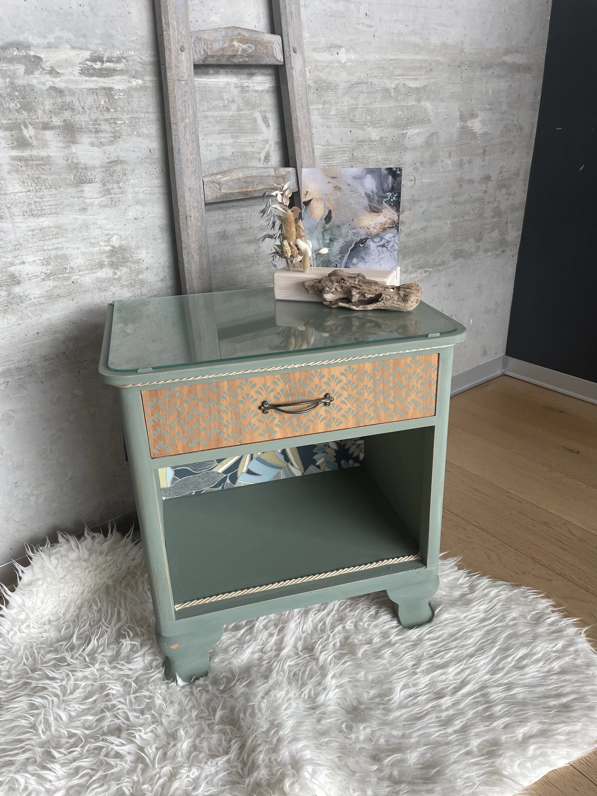 Petite table ancienne vert pastel avec tiroir décoré d'un motif orange et or, décorée de plantes, papier peint floral, dans une pièce avec un mur en béton et un tapis en peluche blanche.