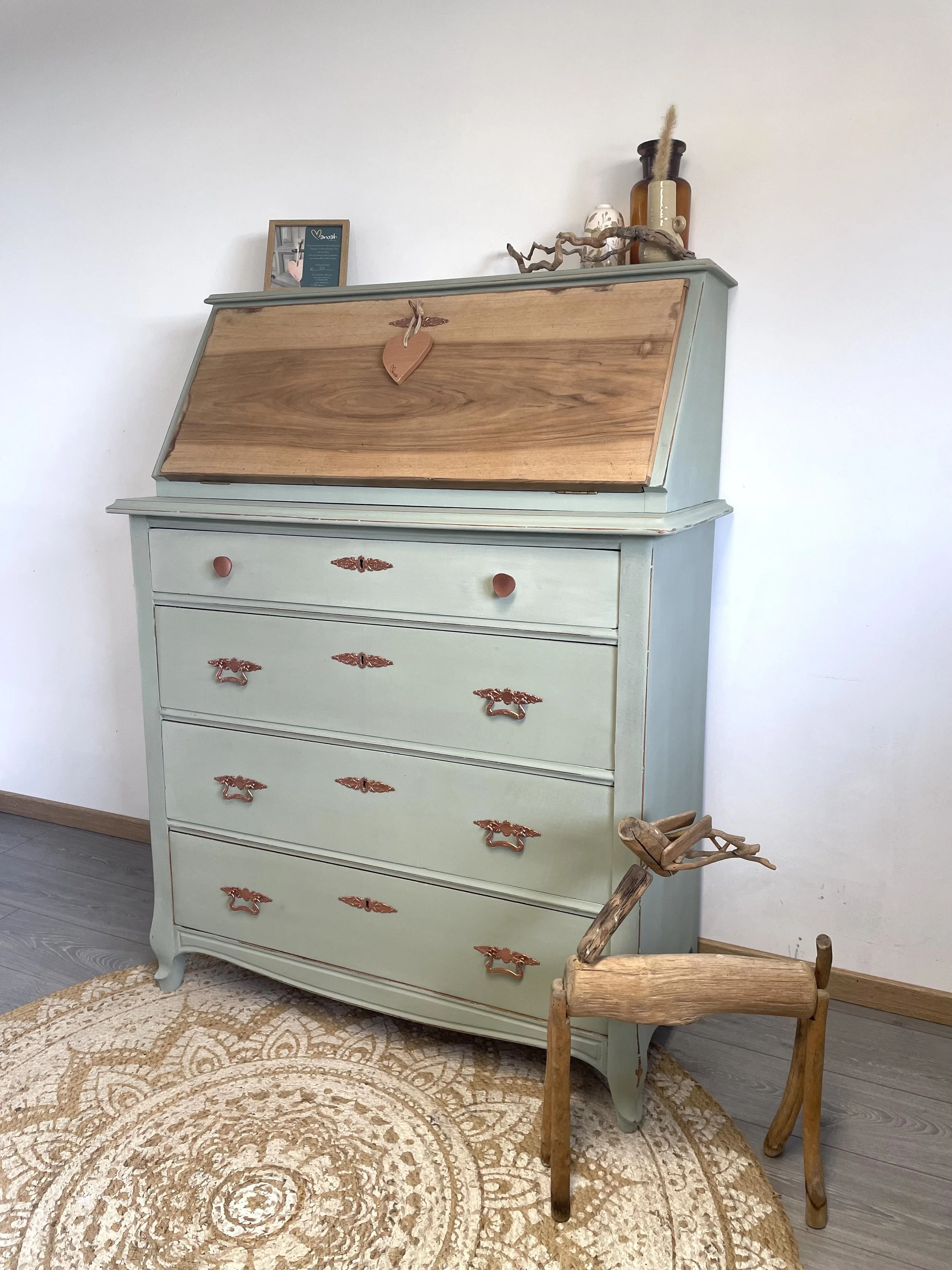 Une commode vert pastel avec des tiroirs, un porte-revues en bois sculpté en forme de cheval, et des objets décoratifs sur le dessus, sur un tapis rond beige et un sol en bois.