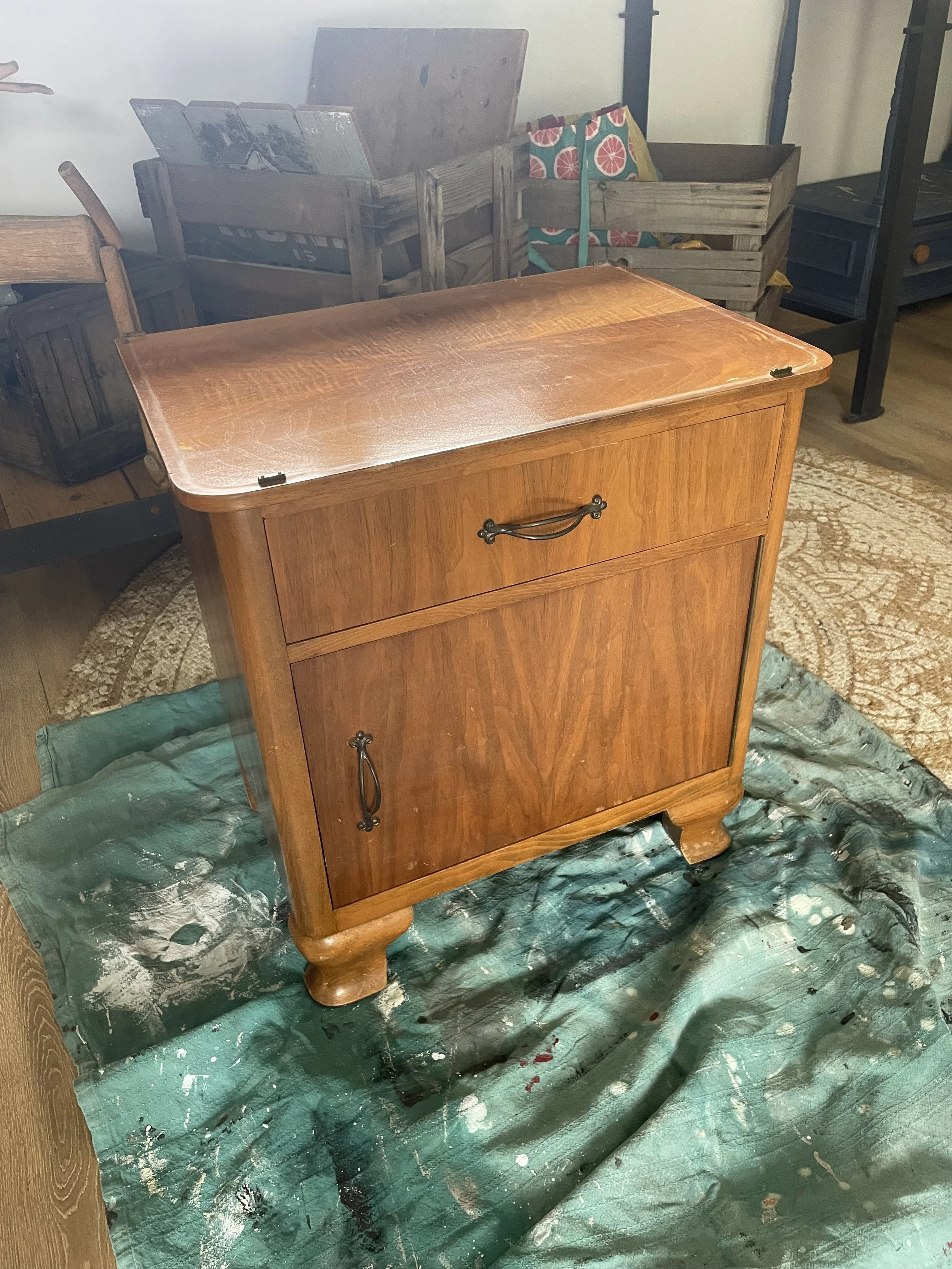 Un meuble en bois ancien avec un tiroir et une porte, placé sur une bâche verte, dans une pièce remplie de caisses en bois et autres meubles.