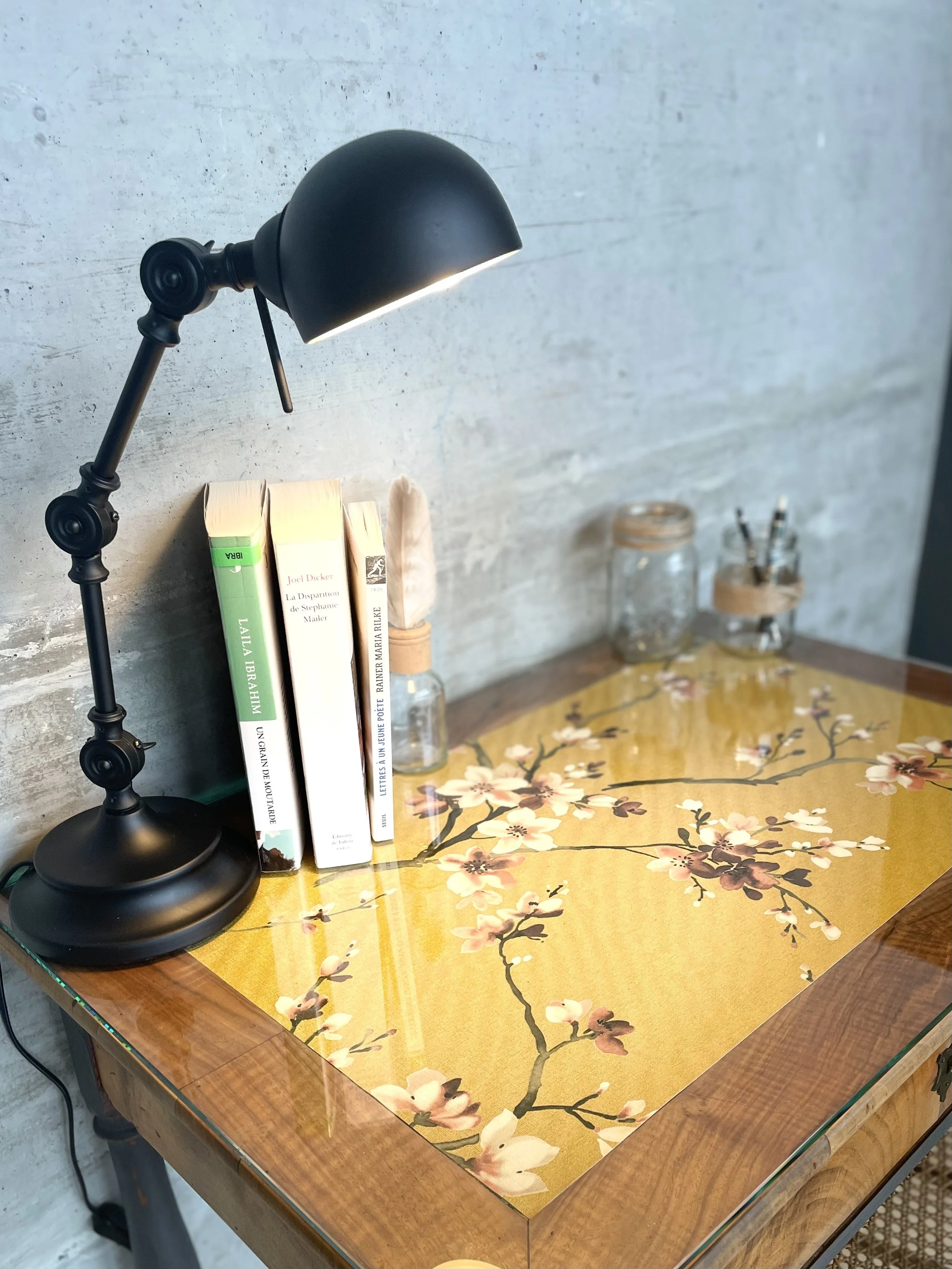 Un bureau avec une lampe de bureau noire, des livres empilés, un bocal à verre, un petit pot en bois avec des stylos et un coton-tige dans un flacon en verre, et une table en bois avec un dessus en verre orné de fleurs de cerisier peintes.