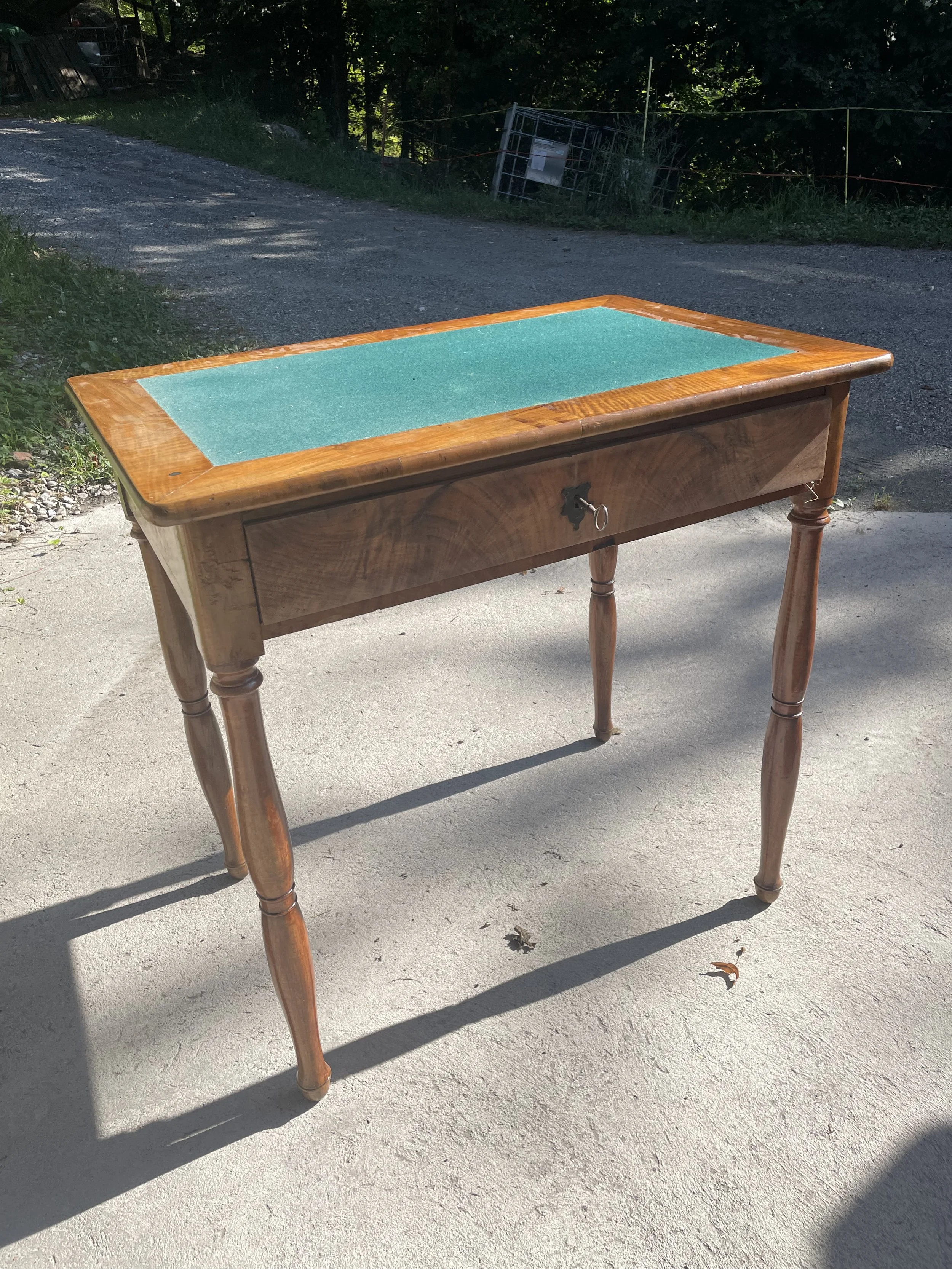 Table en bois avec surface en vert feutre, située à l'extérieur sur un sol en béton, avec des arbres en arrière-plan.