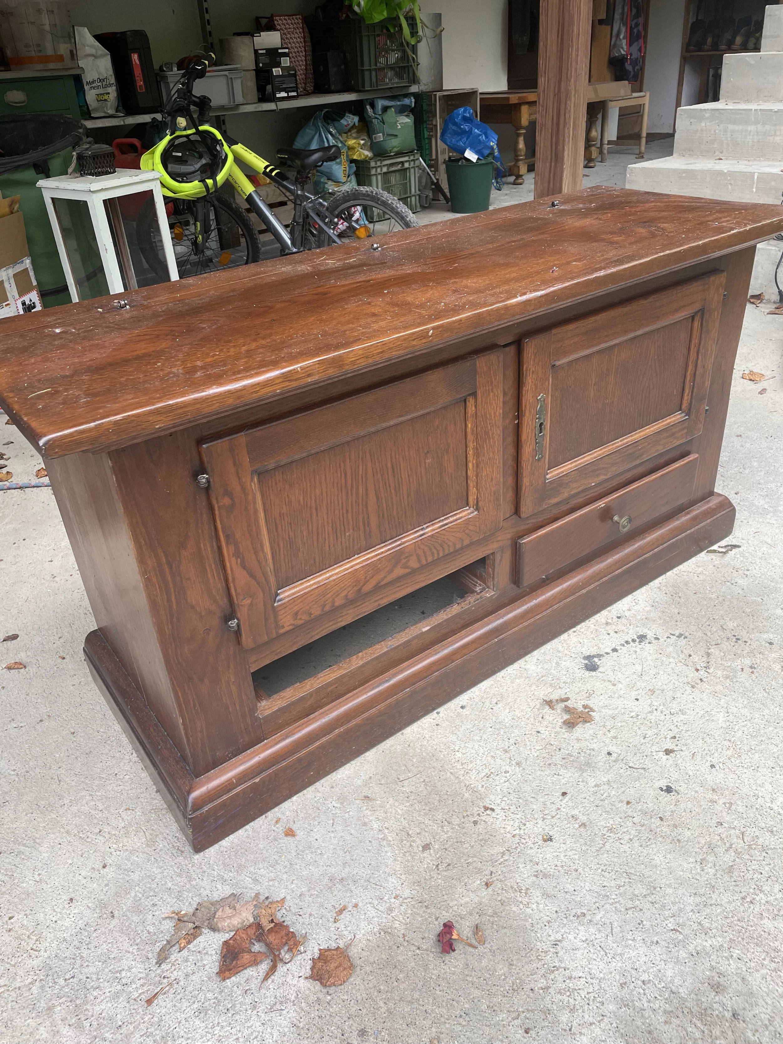 Meuble en bois ancien, probablement un buffet ou un meuble TV, avec deux portes et un tiroir, placé à l'extérieur sur un sol en béton.