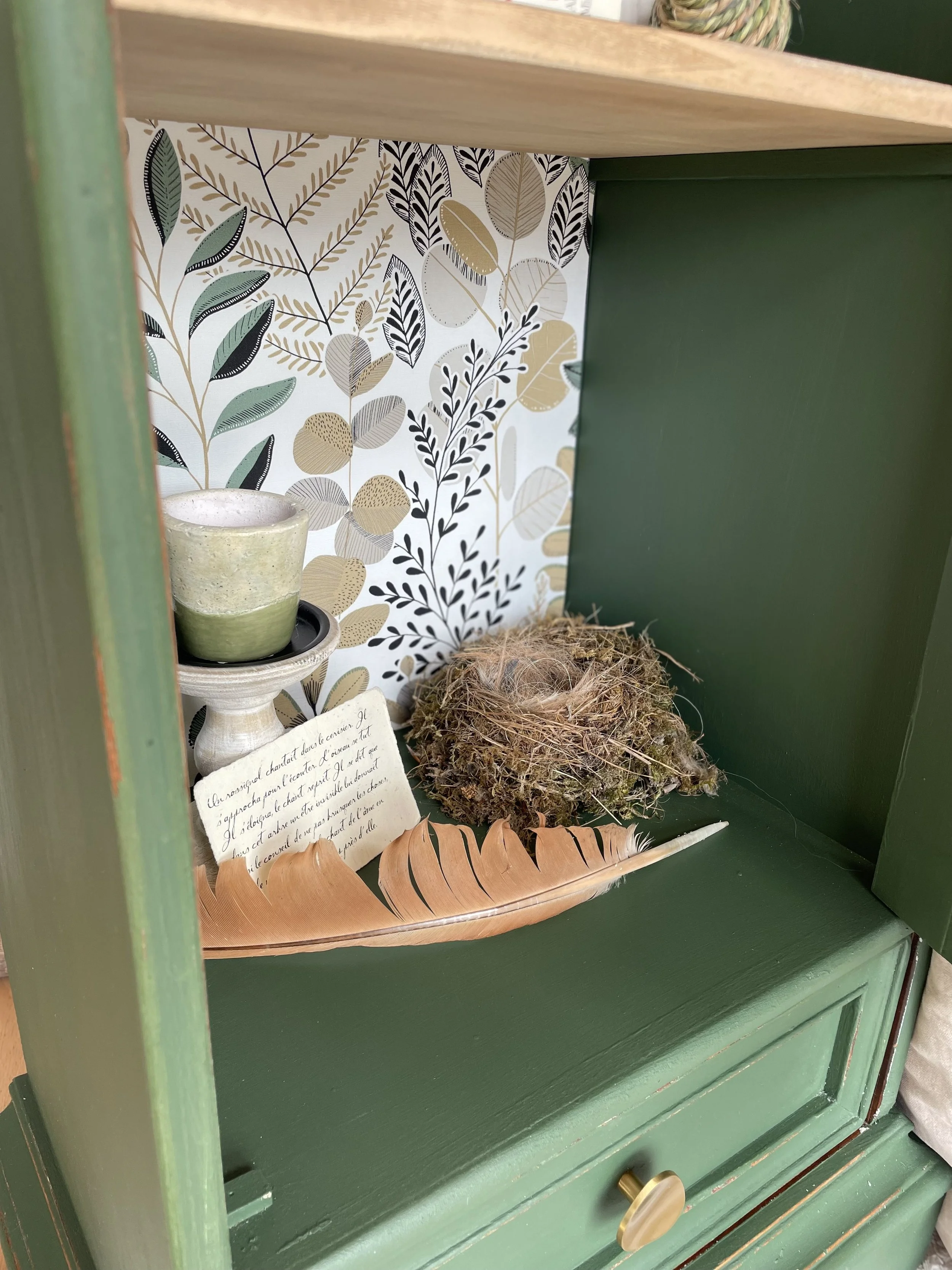Un nid d'oiseaux en mousse, une feuille en plumer, une tasse en céramique émaillée verte sur un support en pierre, un petit carnet avec écritures en français, une scène décorative avec un fond de papier peint à motifs de feuilles. Le tout placé dans une armoire en bois peint en vert.