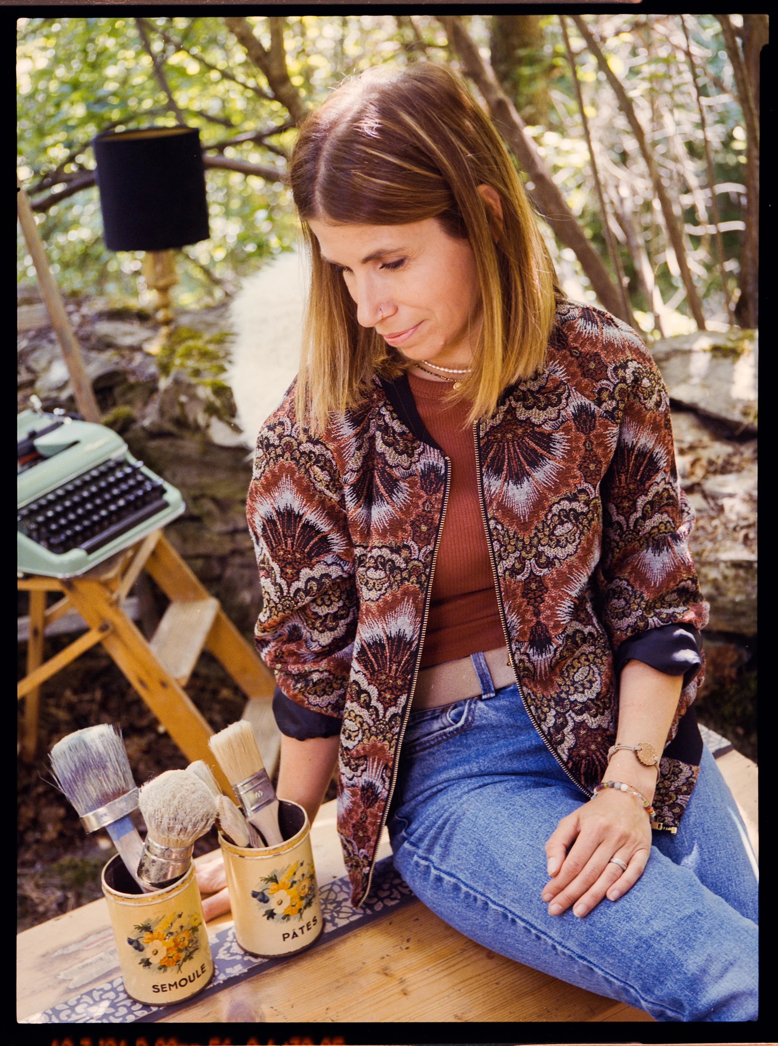 Femme assise dehors, portant un sweat à motifs floraux, avec des pinceaux dans des pots sur une table en bois, dans un environnement naturel.
