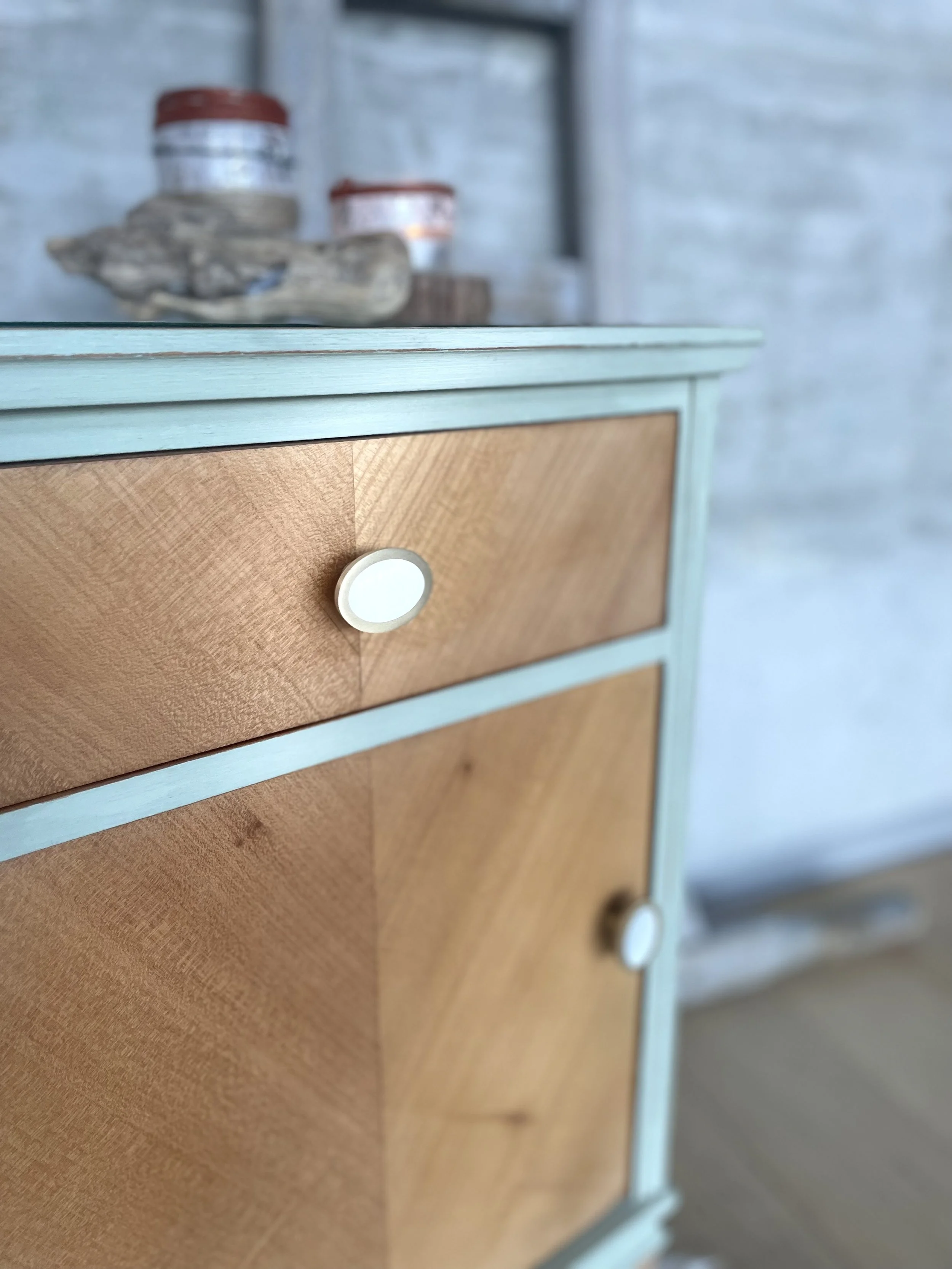 Meuble en bois avec cadre bleu clair, un tiroir en bois clair avec bouton blanc, et une porte en bois clair avec bouton blanc, sur un fond flou avec des boîtes et une pièce de bois sur une étagère.