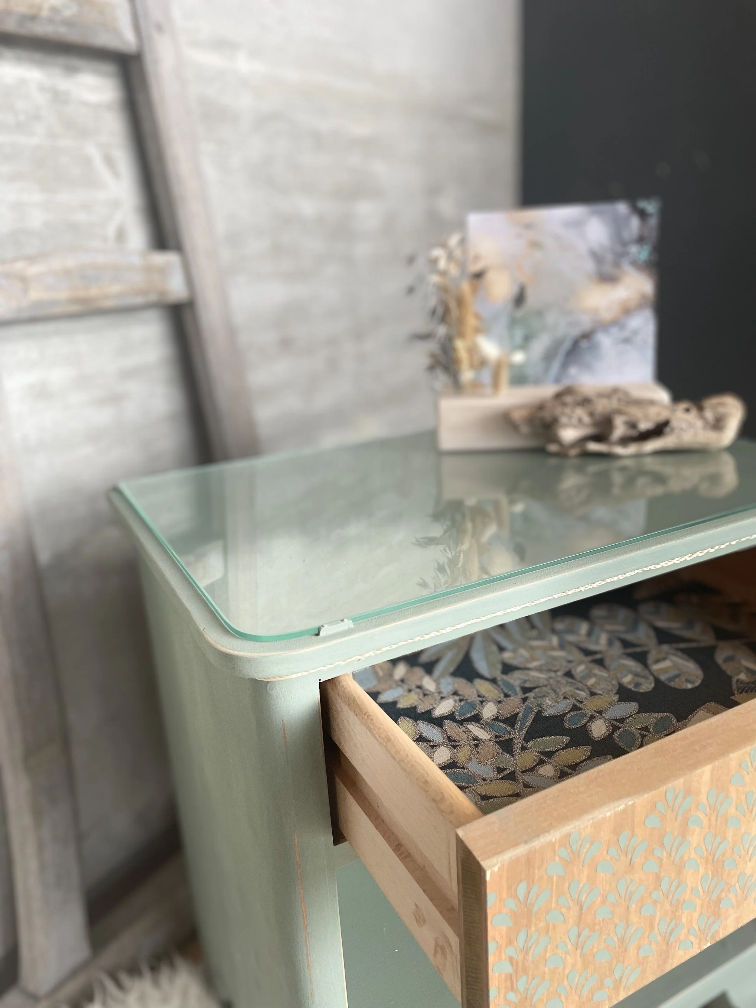 Une table en bois avec un dessus en verre décorée d'un motif floral, placée à côté d'un mur en bois en arrière-plan, avec des décorations et une photo en fond.