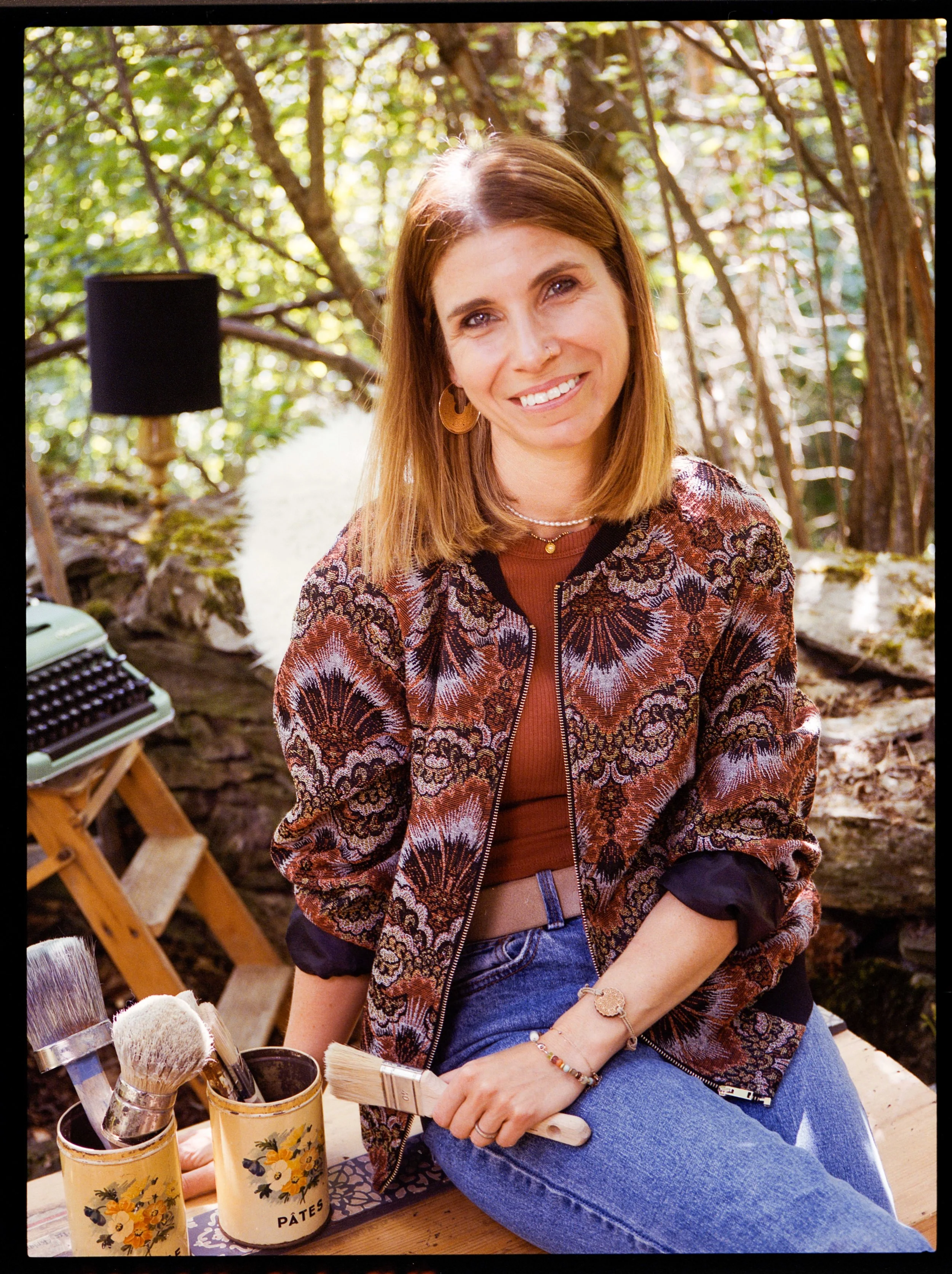 Une femme souriante assise dans un environnement naturel, probablement dans la forêt, avec des outils de peinture ou de maquillage posés devant elle.