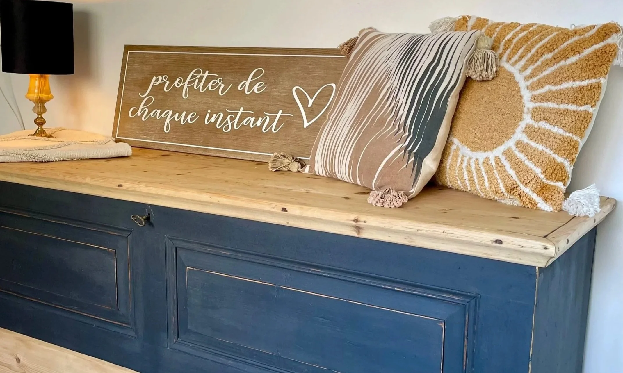 Un meuble en bois avec deux coussins décoratifs, une lampe à côté, et une pancarte en bois avec l'inscription 'profiter de chaque instant' et un cœur dessiné.
