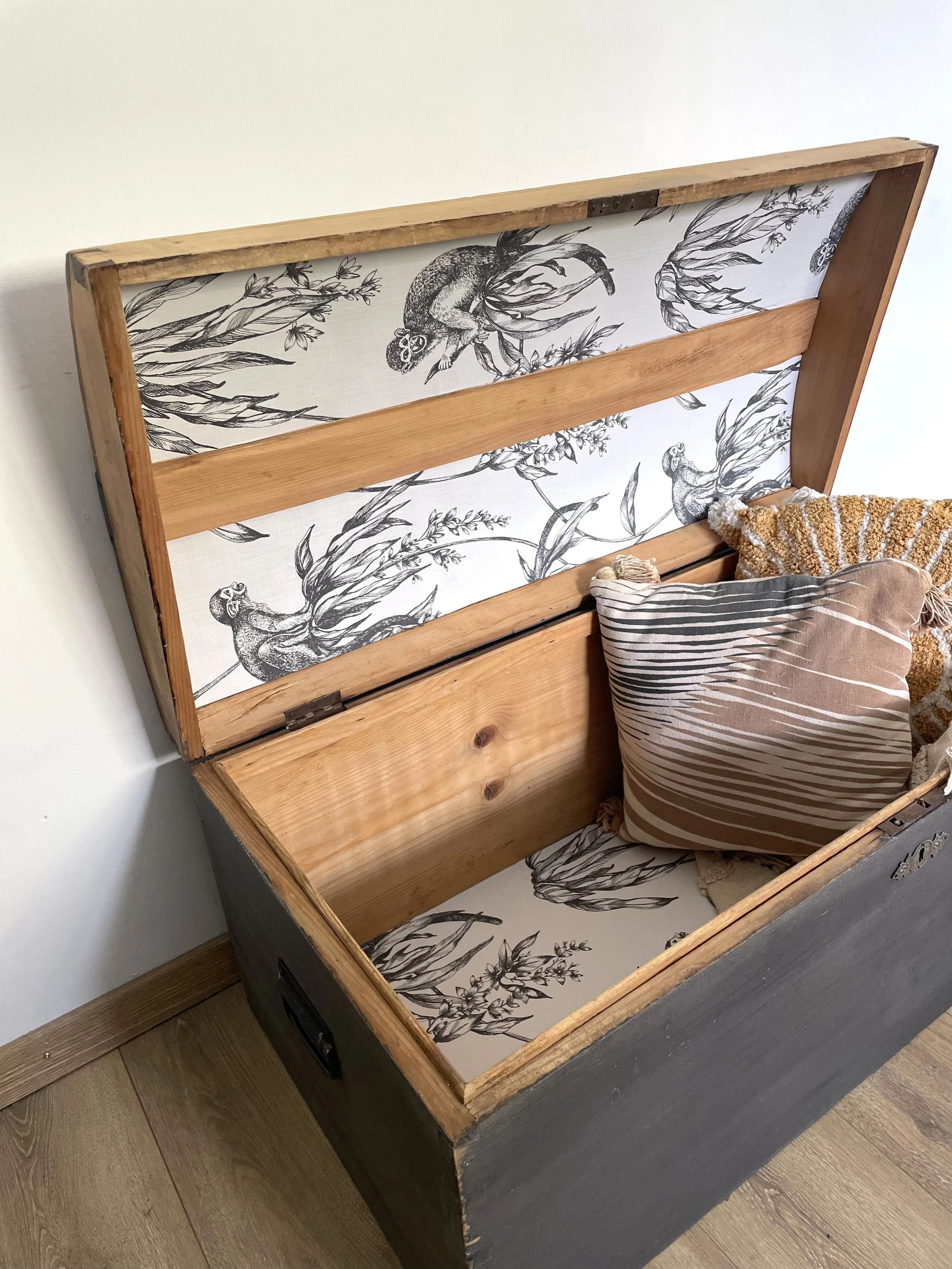 Coffre en bois ouvert avec des coussins à l'intérieur, intérieur recouvert d'un tissu imprimé de dessins de singes et de feuillage.