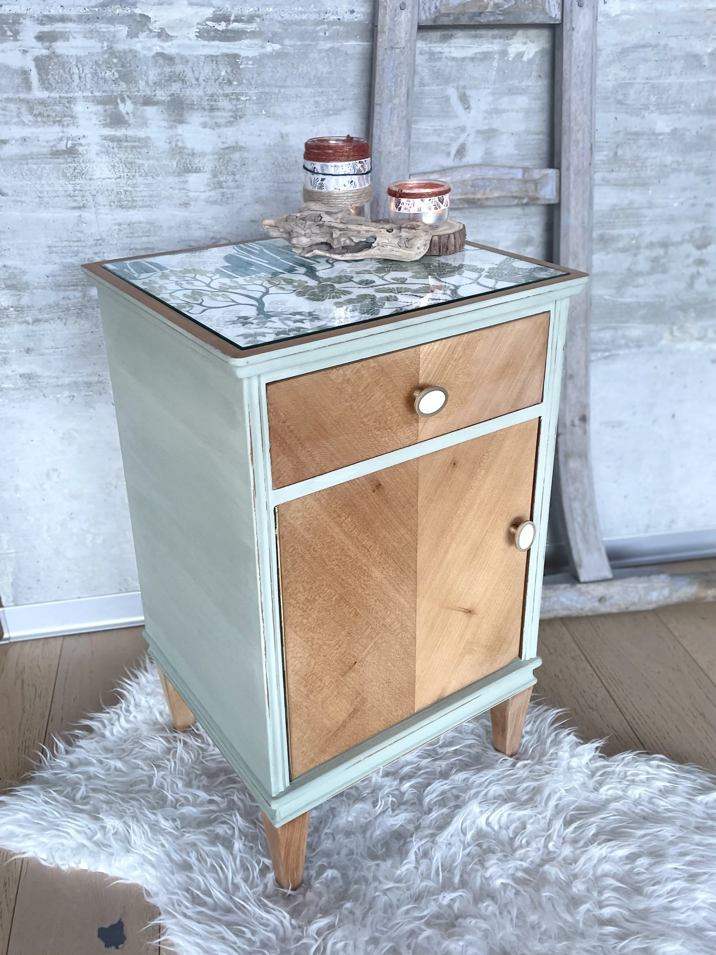 Coffre en bois peint en vert menthe avec un dessus en mosaïque, deux tiroirs avec poignées rondes, décoré avec des bougies et un morceau de bois flotté, posé sur un tapis blanc en peluche devant un mur en béton et une vieille échelle en bois.
