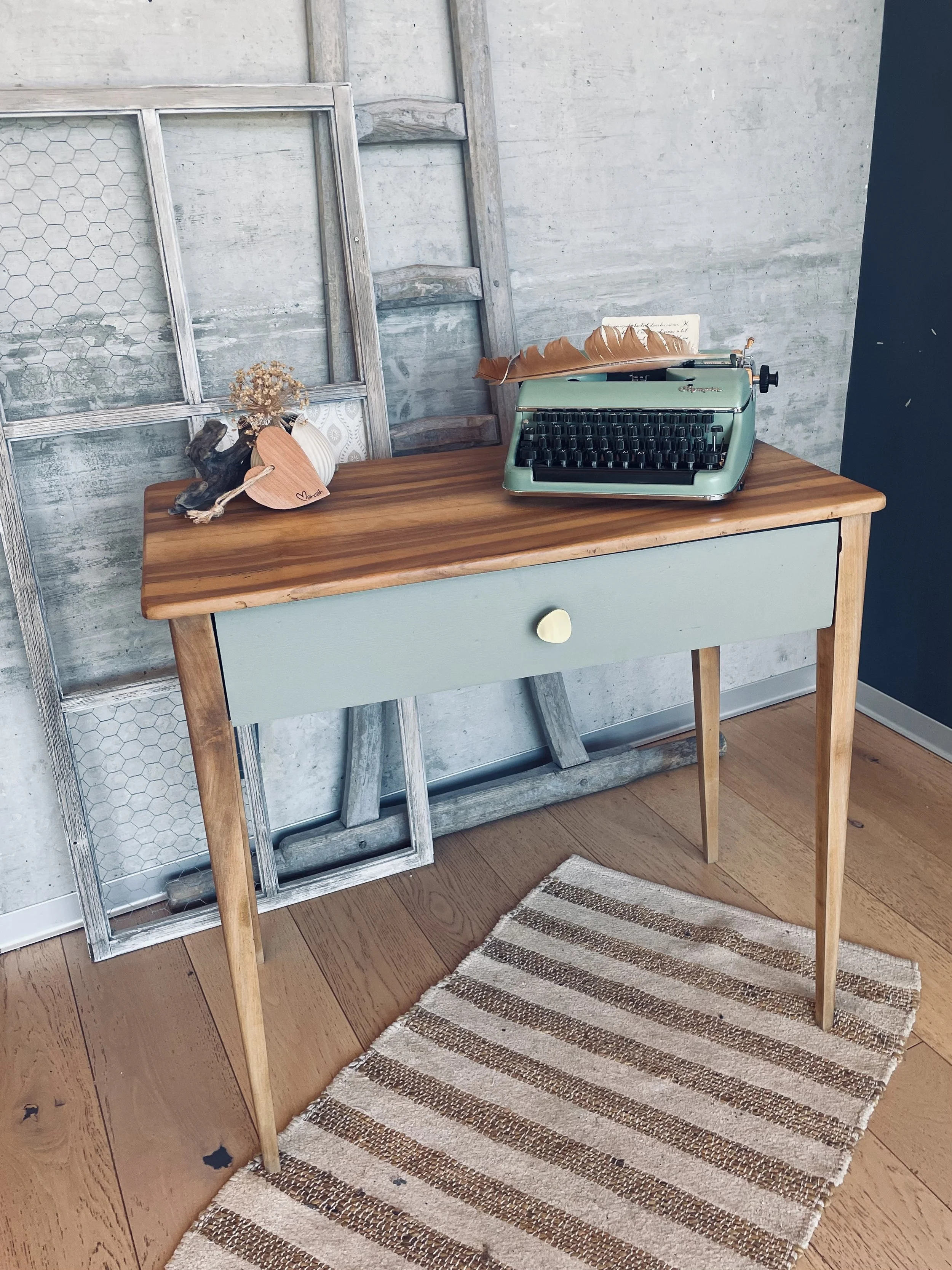 Une table en bois avec un tiroir vert clair et une poignée en forme de cœur, une machine à écrire vintage vert pastel, une décoration avec une branche morte, une petite boule en coton, un cadre en bois et un tapis rayé beige et marron sur un sol en bois.