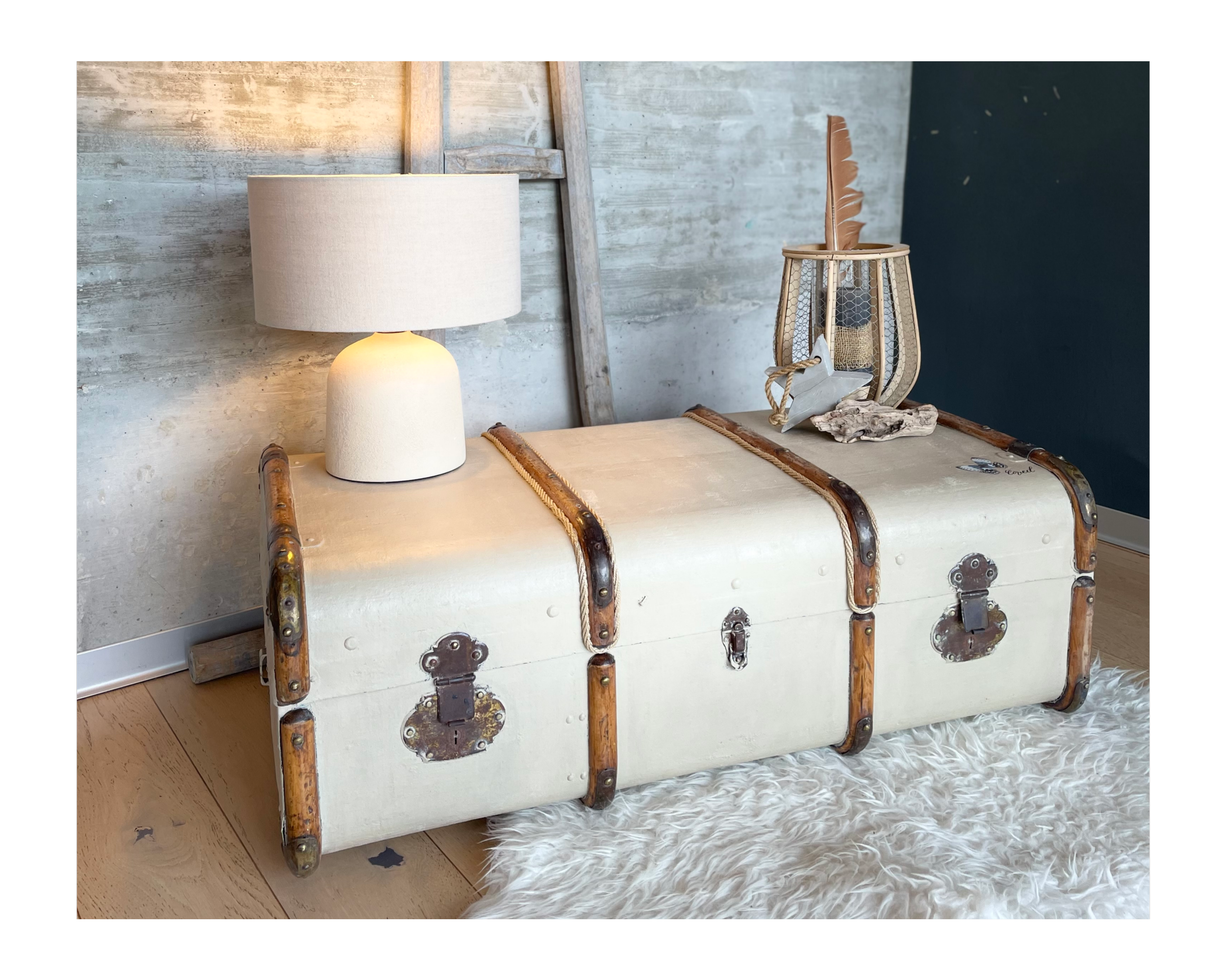 Une morse en céramique décorative posée sur un coffre beige avec ferrures en métal, flanqué d'une lampe de table blanche et d'un vase en rotin avec une plume en décoration, sur un tapis en laine blanche