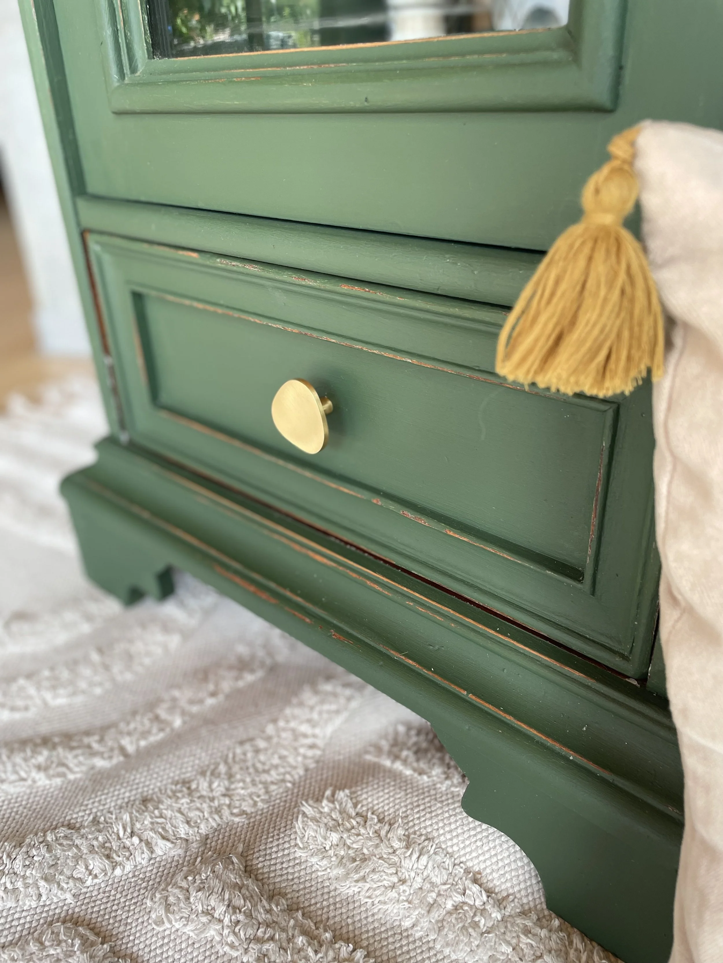 Meuble vert avec un tiroir, une poignée en forme de cœur en métal doré, et une étoffe blanche avec une pompon jaune à droite, posé sur un tapis tissé beige.