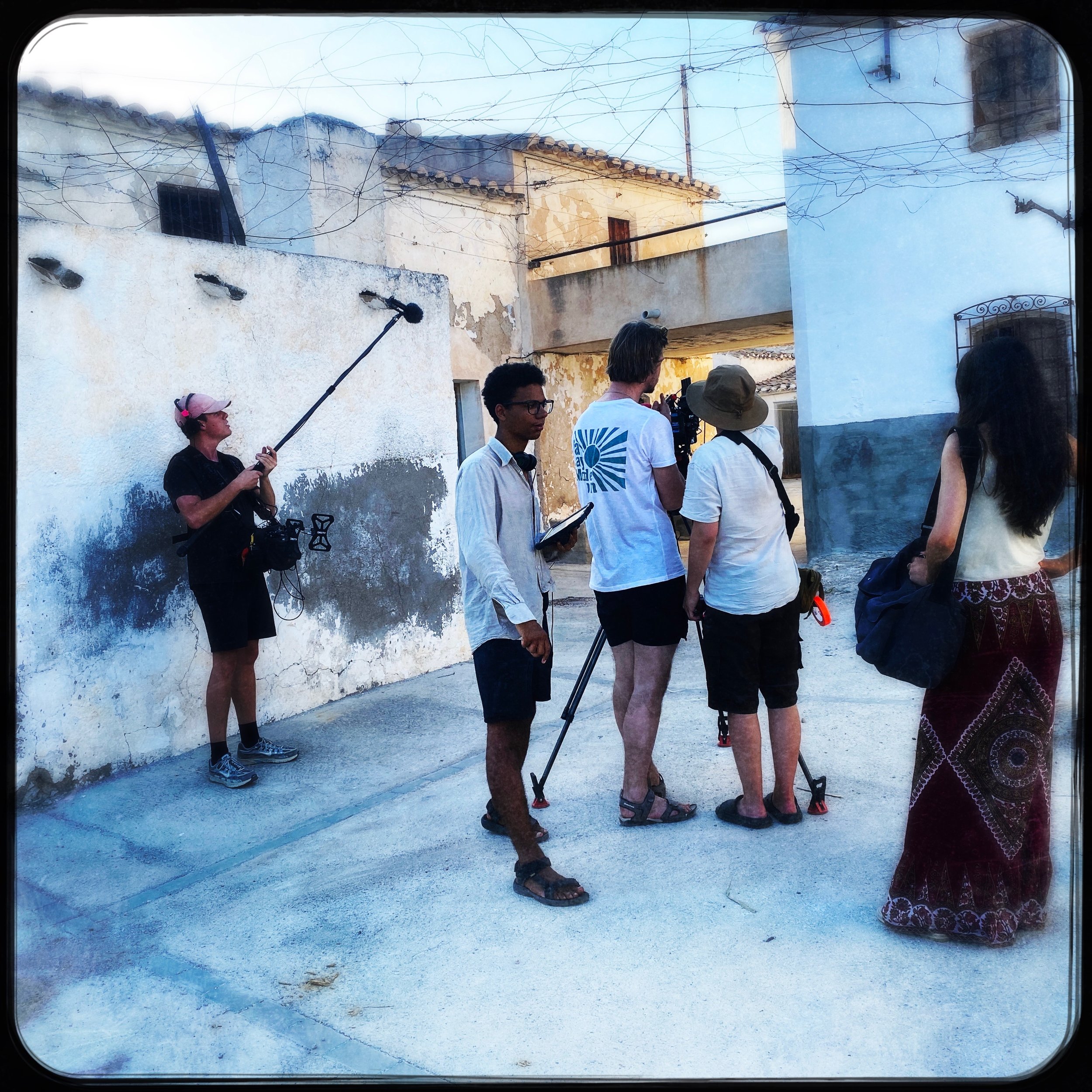 A group of five people on a film set outdoors, with a woman holding a boom microphone, crew members with equipment, and a woman in a bohemian skirt standing to the right, against a backdrop of rustic buildings with weathered walls.