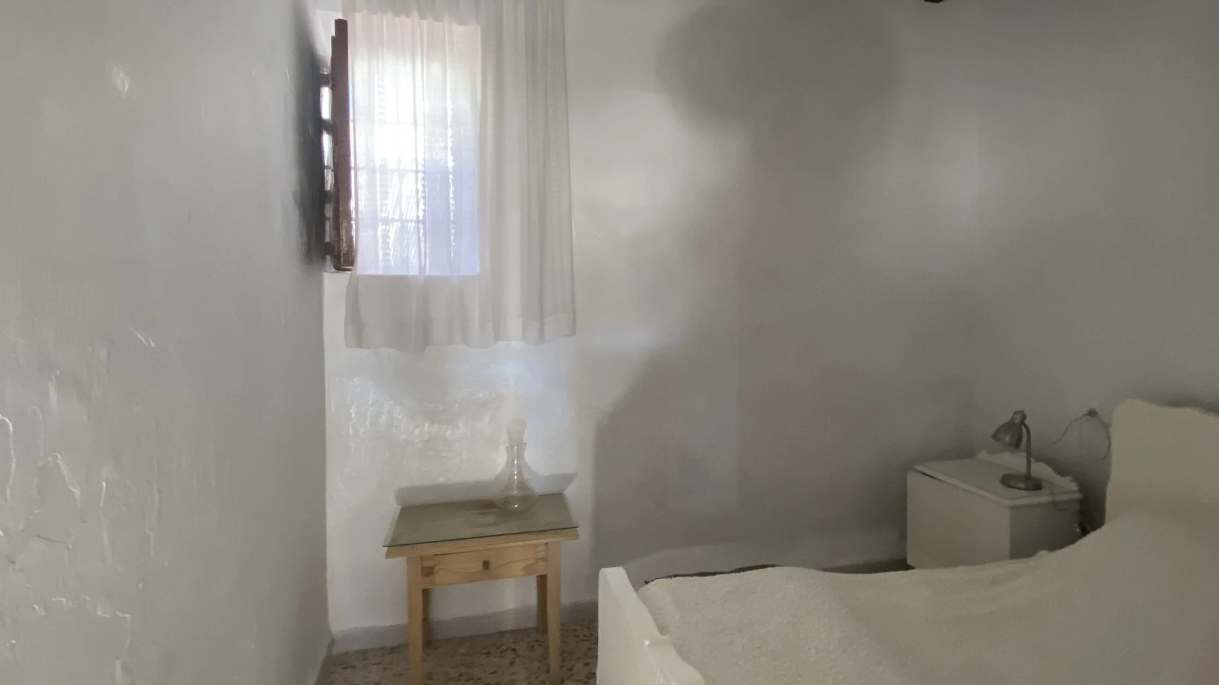 A minimal bedroom with white walls, a bed with a white headboard and blankets, a small white nightstand with a lamp, a wooden side table with a glass vase, and a window with sheer curtains letting in light.