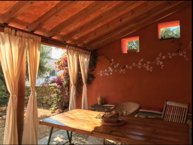 Sunlit patio with wooden table, wicker chairs, and cream curtains, surrounded by plants and small windows in a warm orange wall.