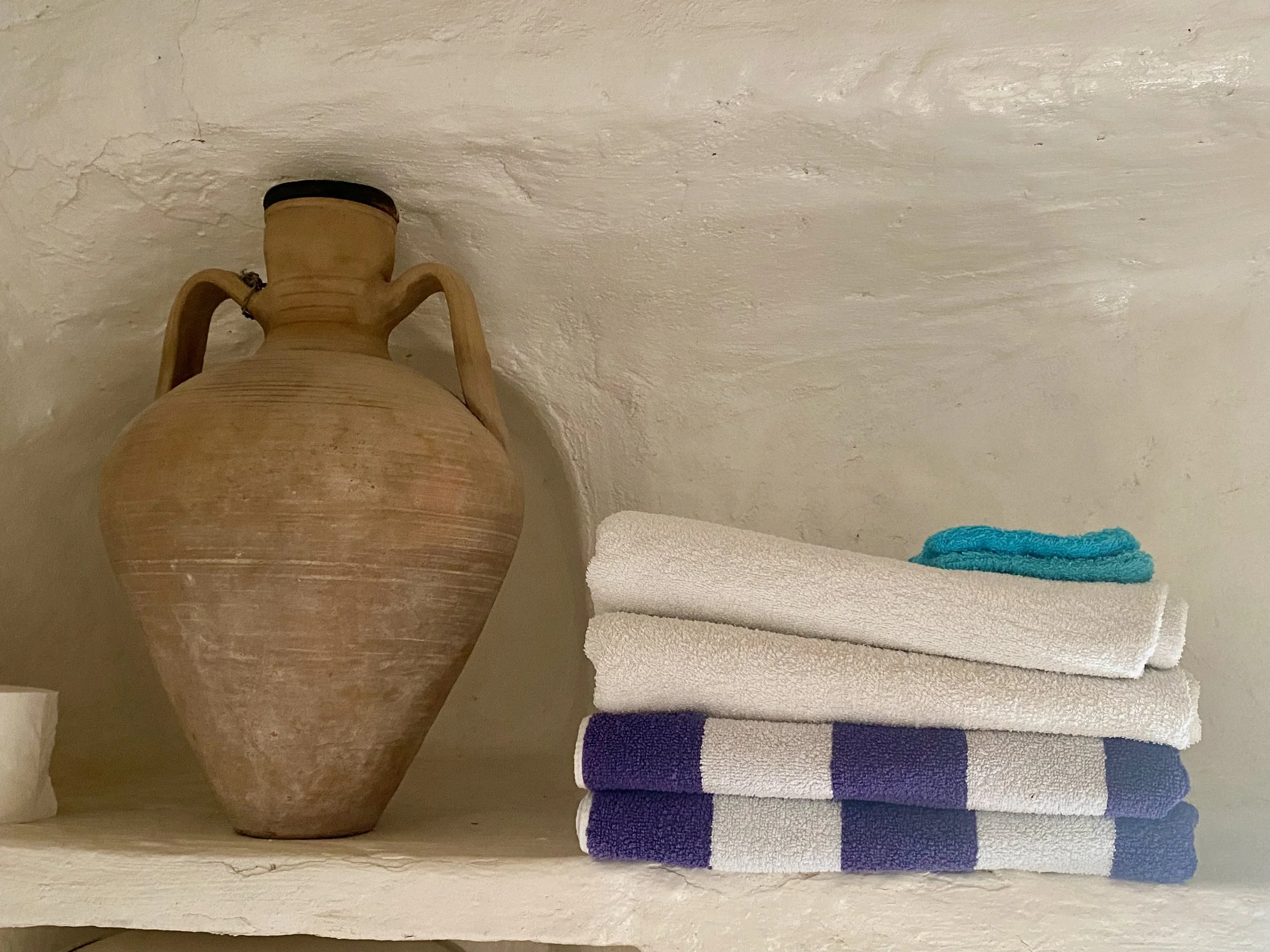 A large, decorative ceramic vase with handles placed on a cream-colored surface, next to a neatly folded stack of three white towels, and a small blue towel on top, against a textured white wall background.