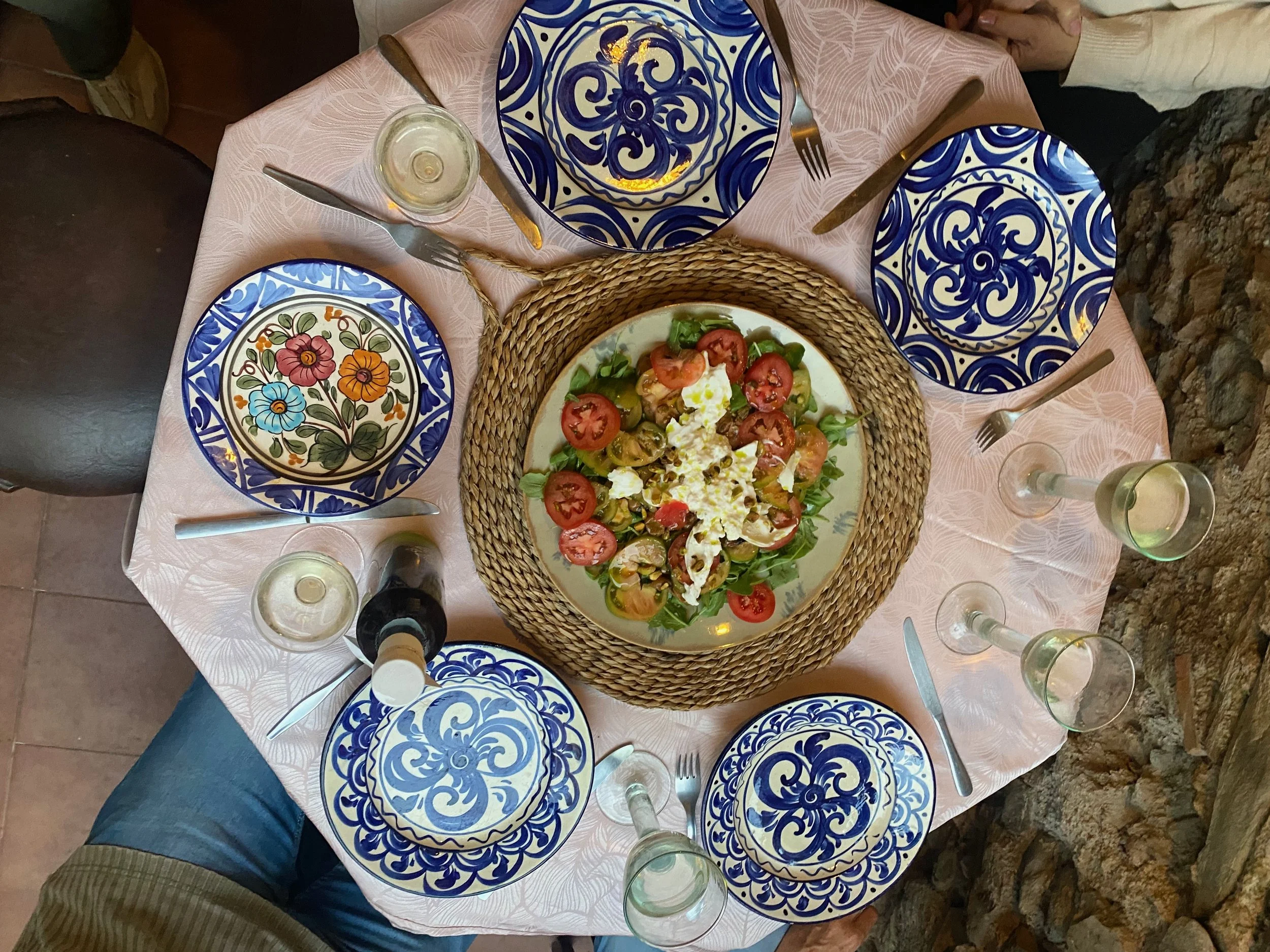 A round table set for a meal with five colorful, patterned plates, four glasses of water, and a bottle of white wine. In the center is a bowl of mixed salad with tomatoes, lettuce, and cheese, surrounded by a woven placemat.