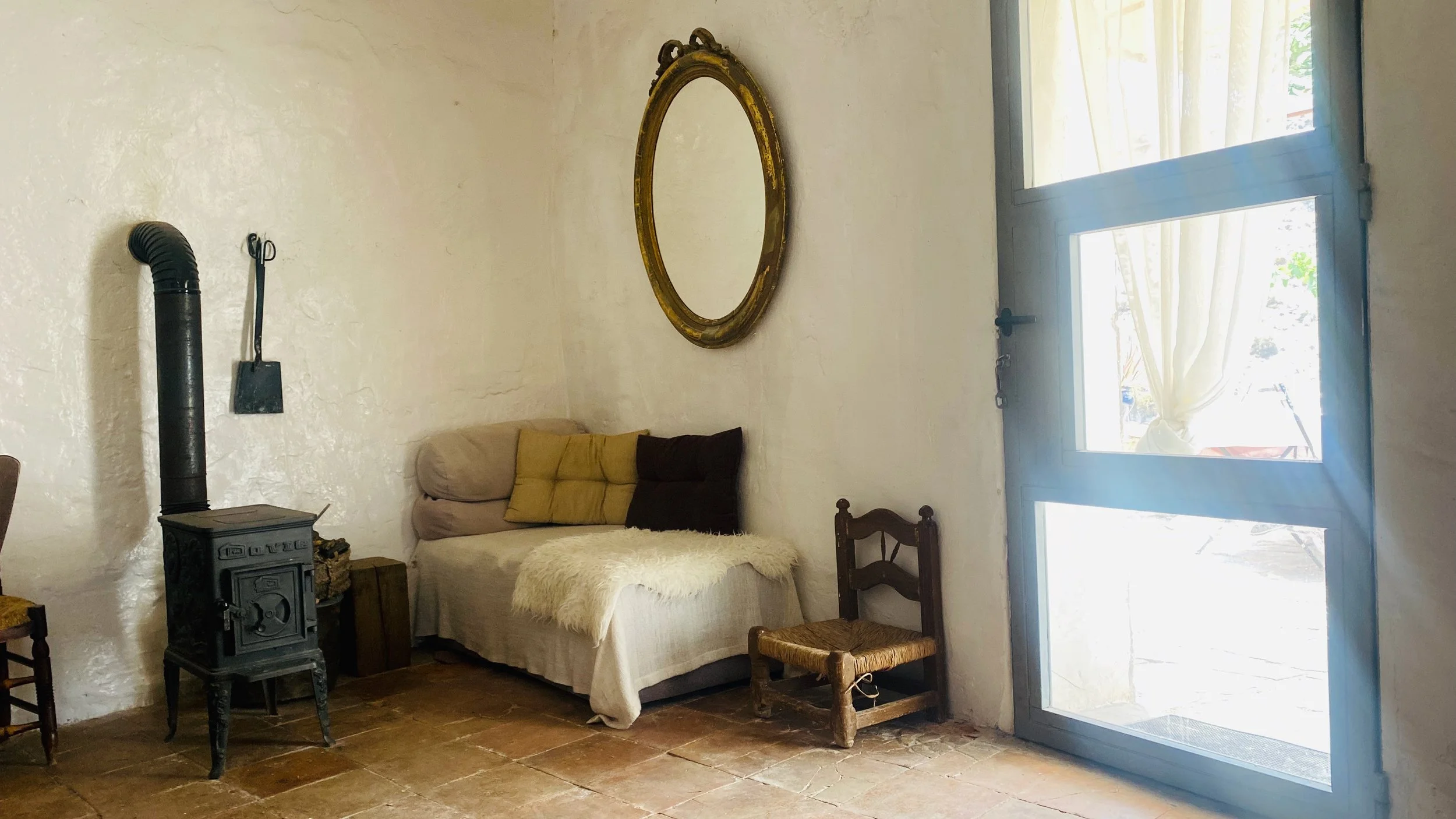 Cozy living room with a cream-colored wall, a small wood stove, a plant-themed chair, a beige sofa with pillows, a fluffy throw blanket, an oval mirror, and a large glass door with curtains.