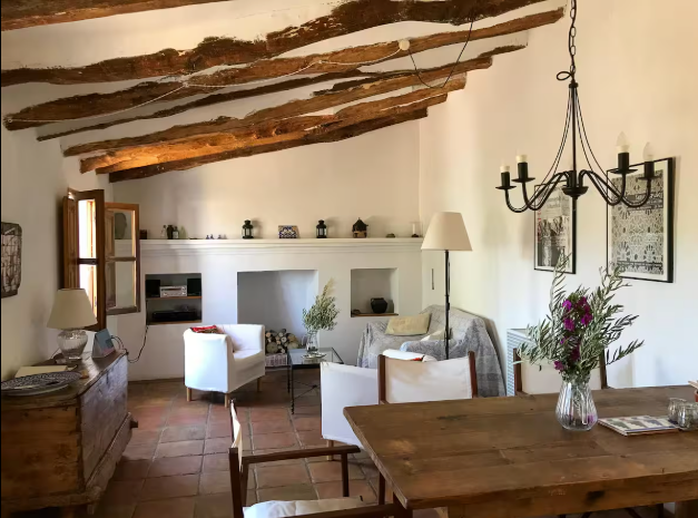 Cozy living room with exposed wooden beams, white walls, a sofa with cushions, chairs, a wooden dining table with a vase of flowers, and decorative elements.